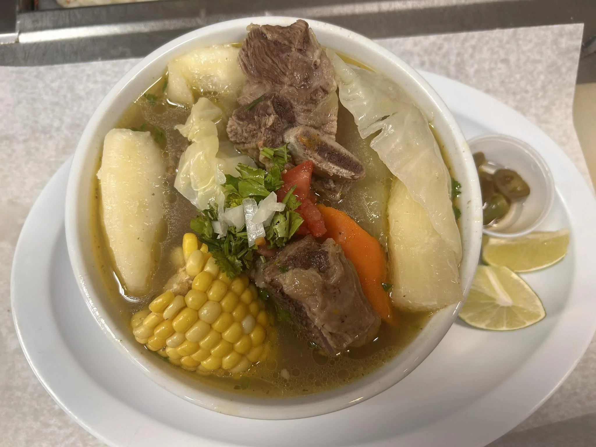 A bowl of beef soup with potatoes, corn, carrots, cabbage, chopped cilantro, and seasonings, served with lemon wedges and lime slices on the side.
