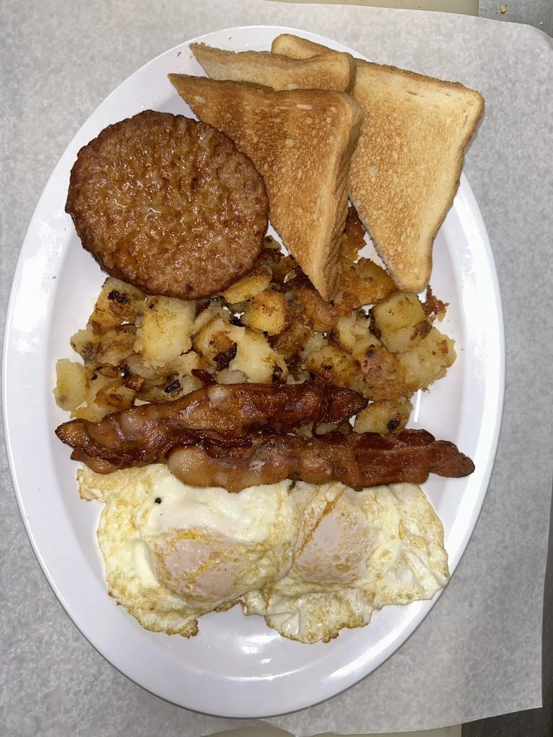 Breakfast plate with fried eggs, bacon, hash browns, toast, and sausage patty.