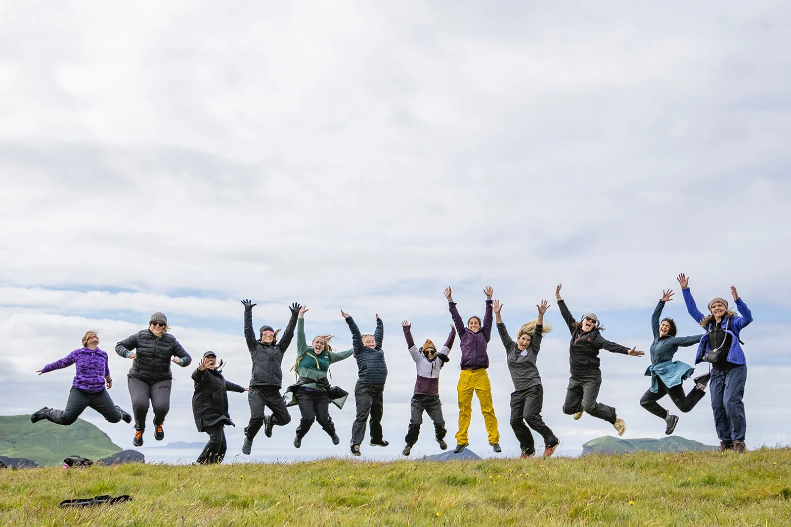 A group of people jumping in the air on a grassy field with cloudy sky and distant mountains.