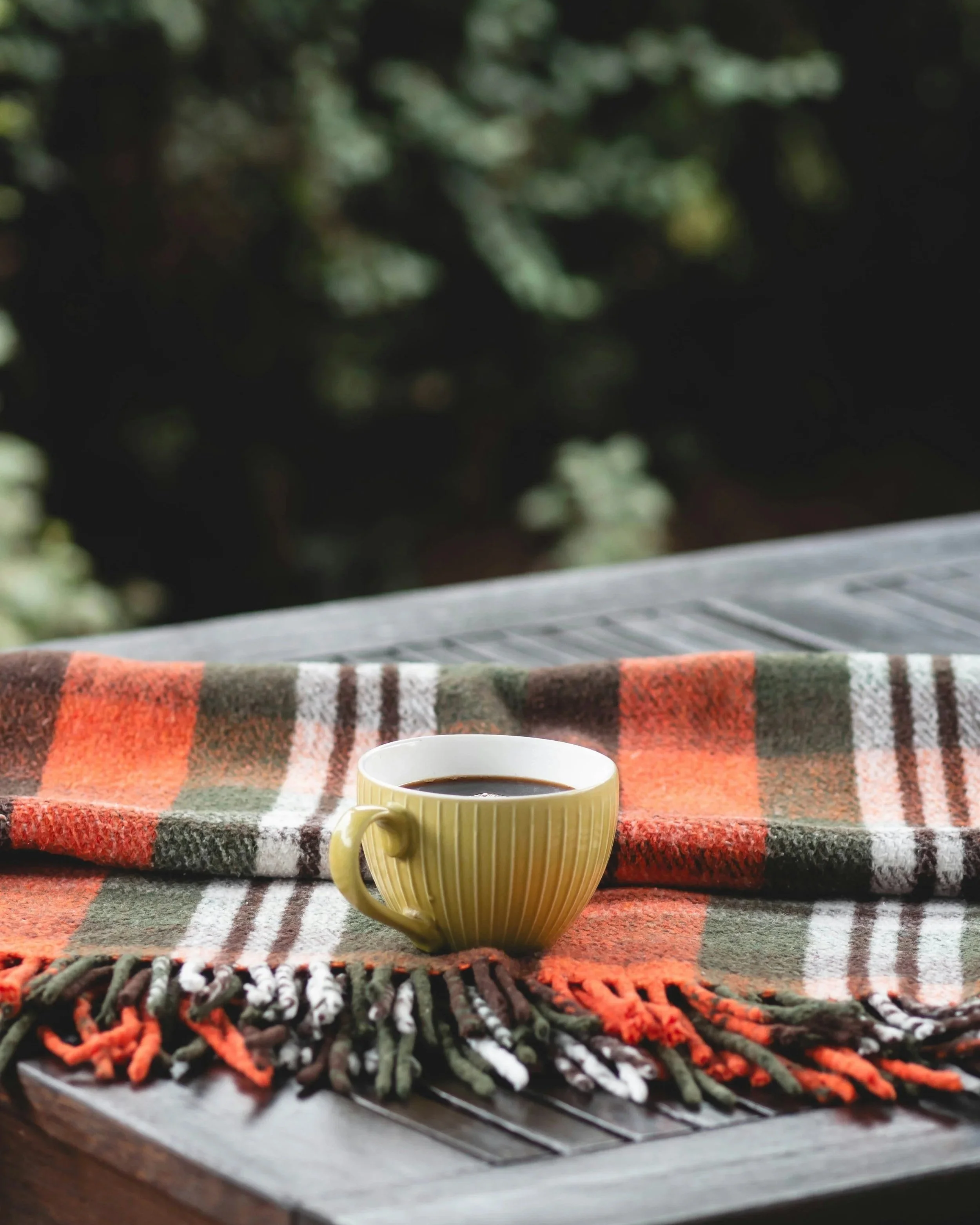 A yellow ceramic mug filled with black coffee placed on a wooden table, with a colorful plaid blanket underneath and a blurred outdoor background of trees.
