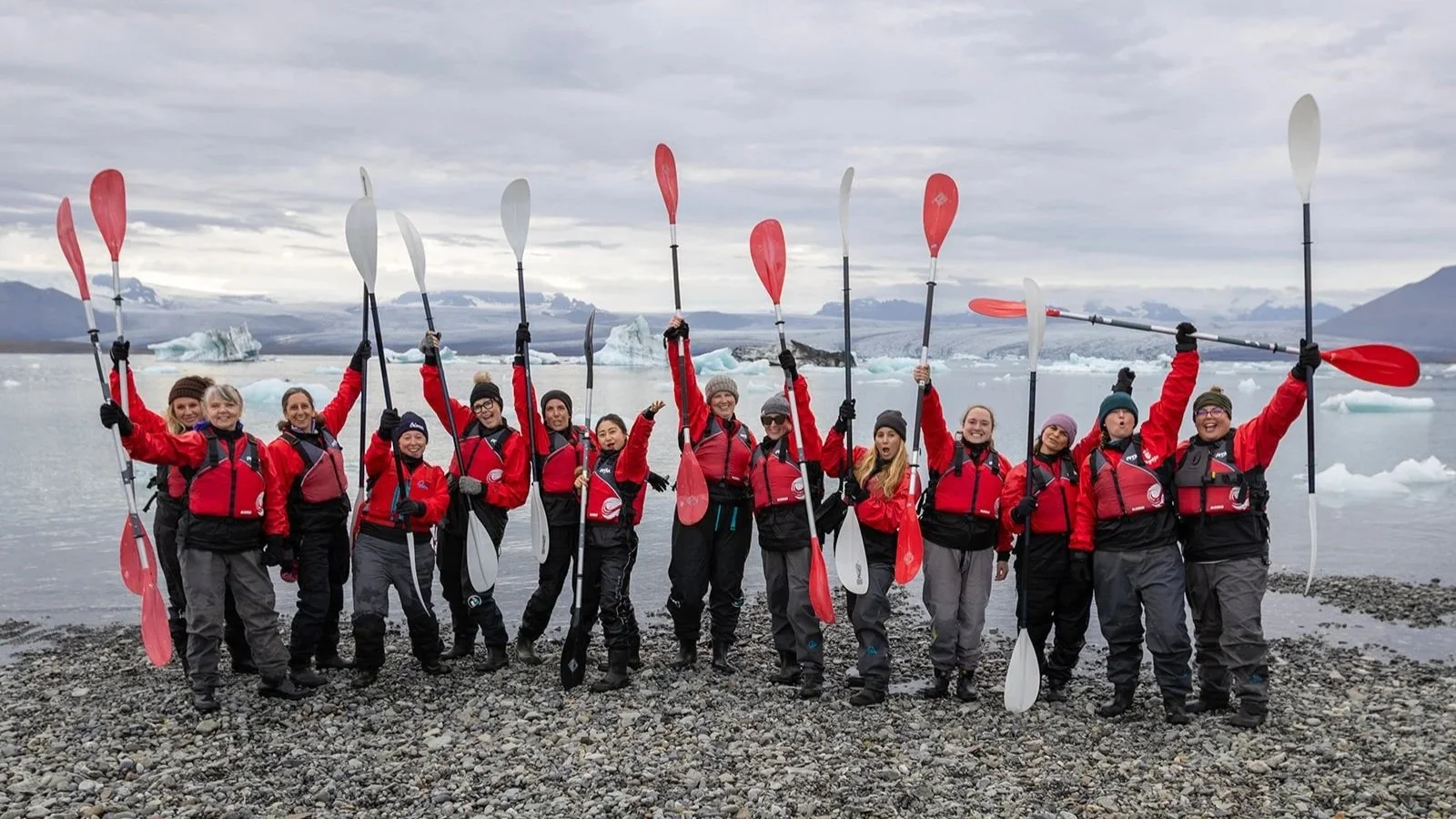 Group of people in red jackets and black pants holding paddles on a rocky shoreline with icebergs and mountains in the background.