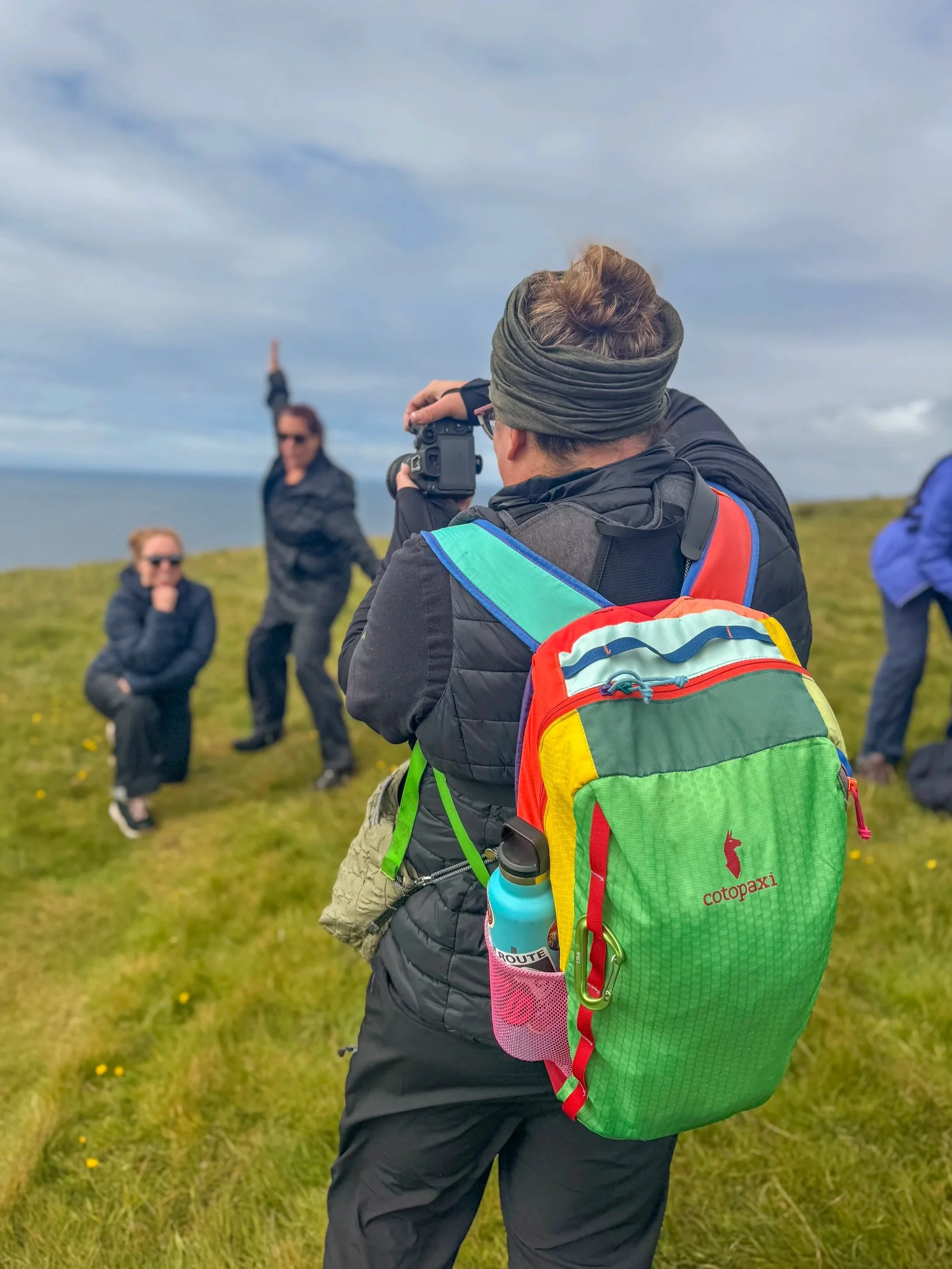 A person taking a photo of three women posing outdoors on a grassy hill with a body of water in the background. The person has a green and red backpack and is wearing a head wrap.