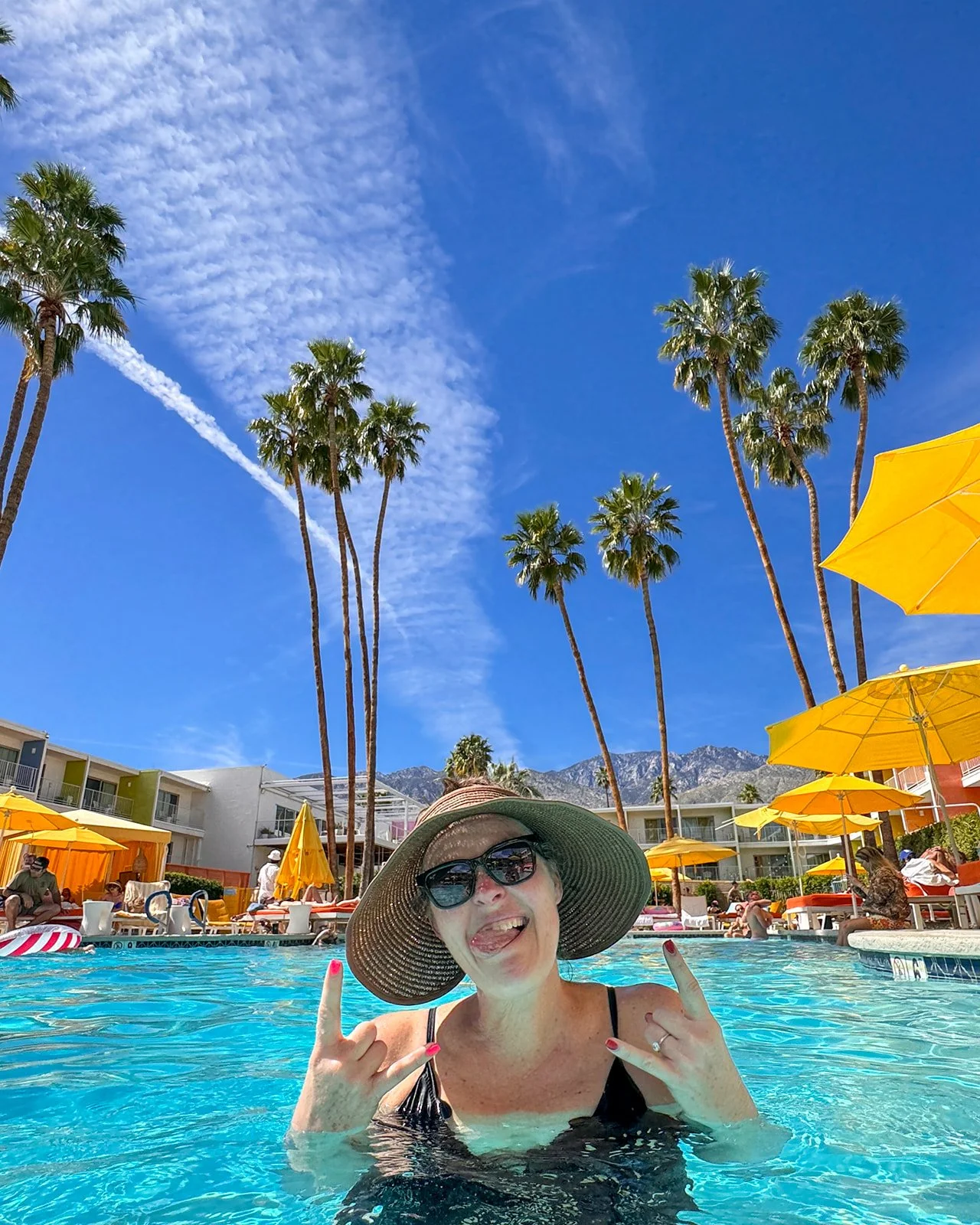 Woman wearing large sun hat and sunglasses in a pool with a smile, showing rock formations and palm trees under a blue sky with clouds and yellow umbrellas in the background.