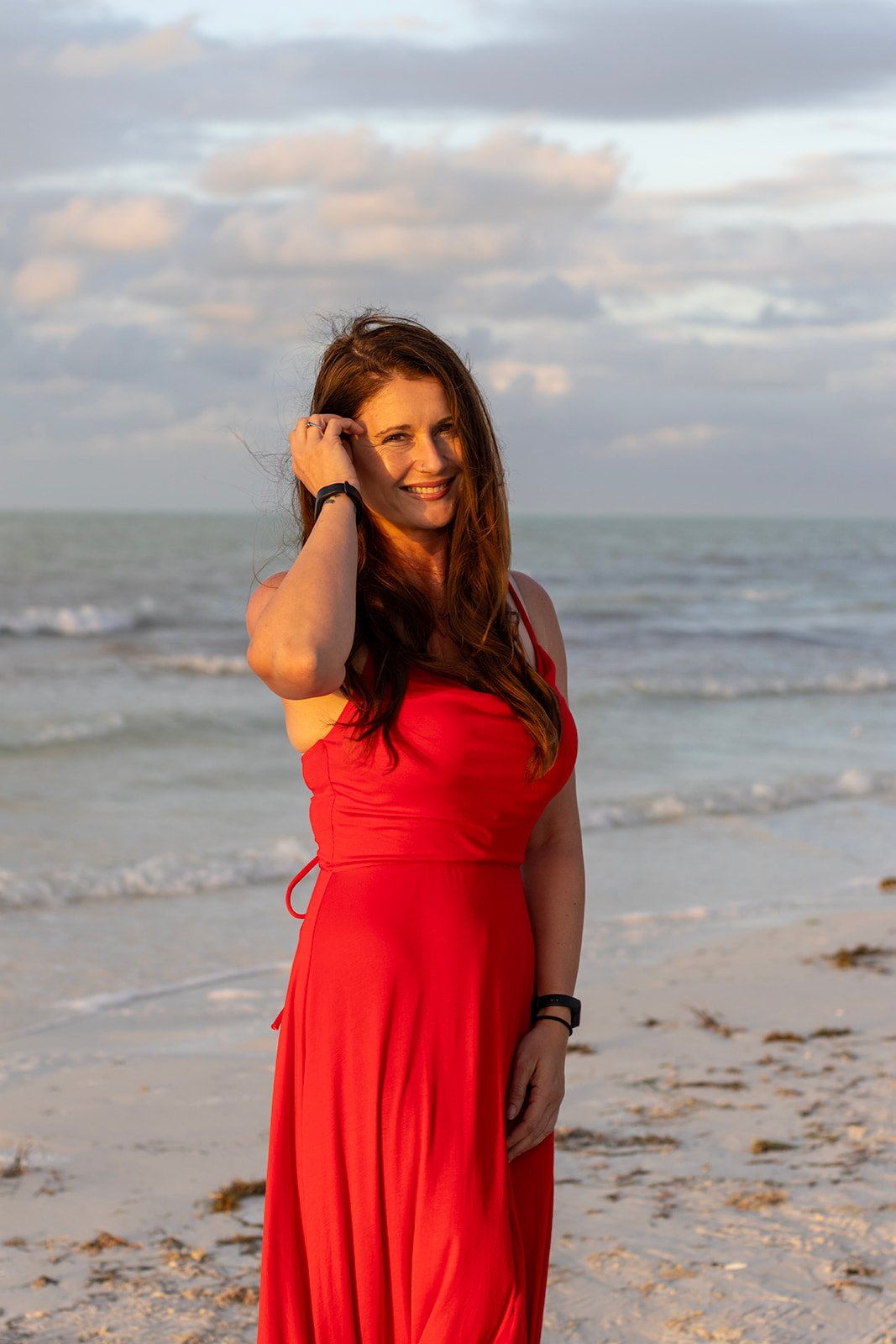 Woman in a red dress standing on a sandy beach with the ocean and cloudy sky in the background, smiling, during sunset.