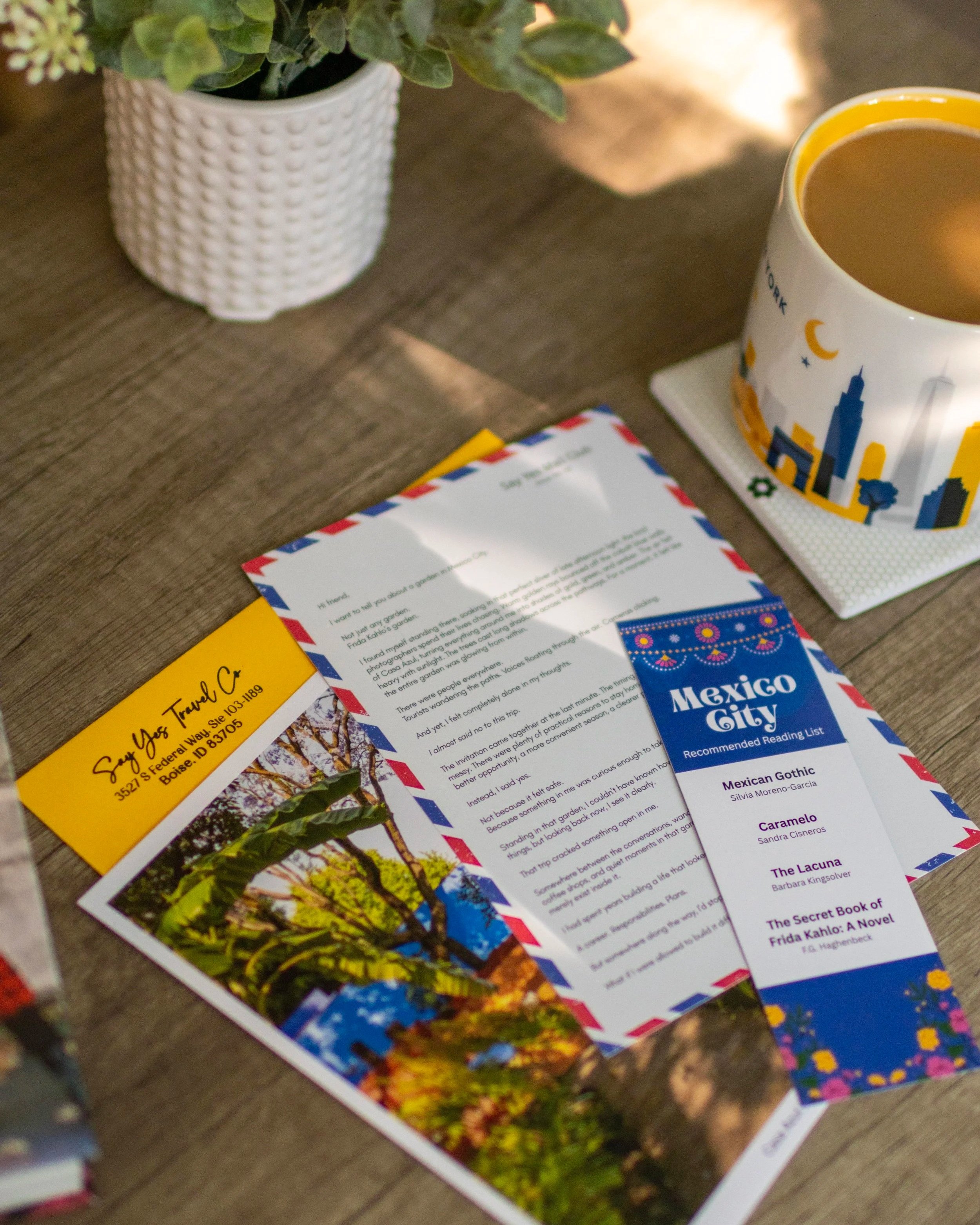 Photograph of a table with a white potted plant, a cup of coffee with a cityscape design, a recommended reading list titled 'Mexico City', a yellow business card, and some colorful paper items under warm lighting.