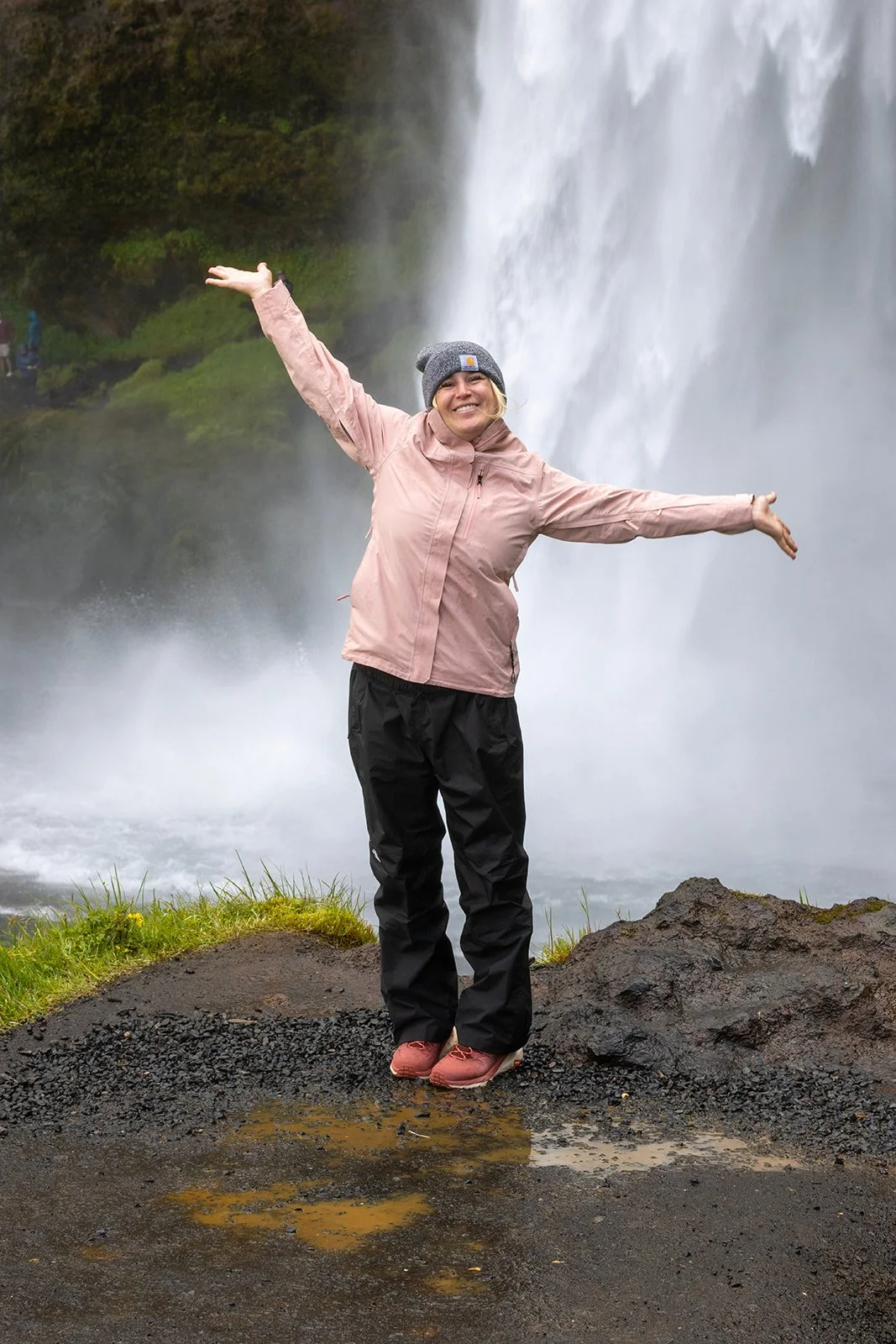 A woman wearing a pink jacket, black pants, and a gray beanie standing with arms outstretched in front of a waterfall.