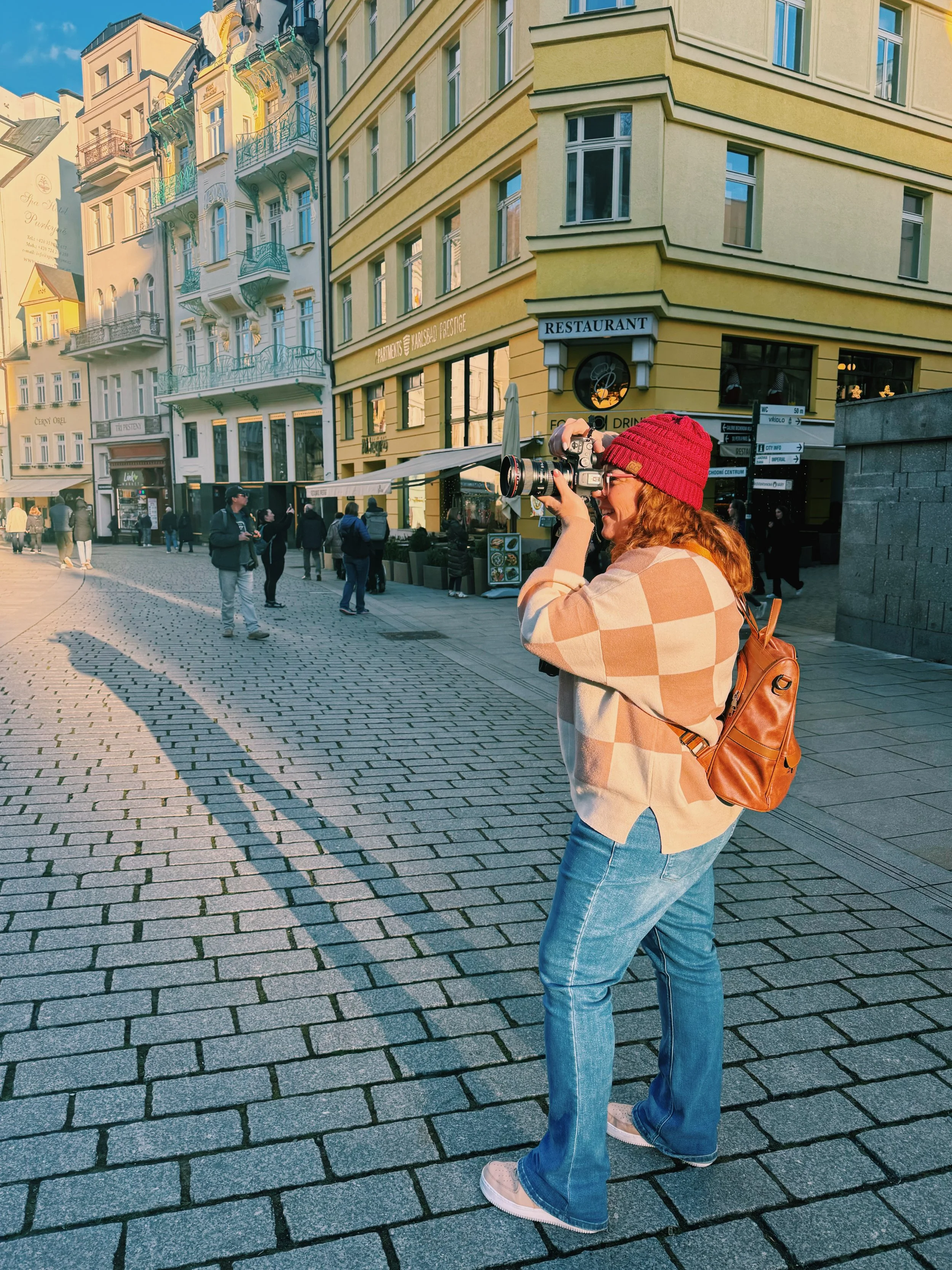 Woman in a red beanie and beige checkered sweater taking photos with a camera on a cobblestone street in a busy city.