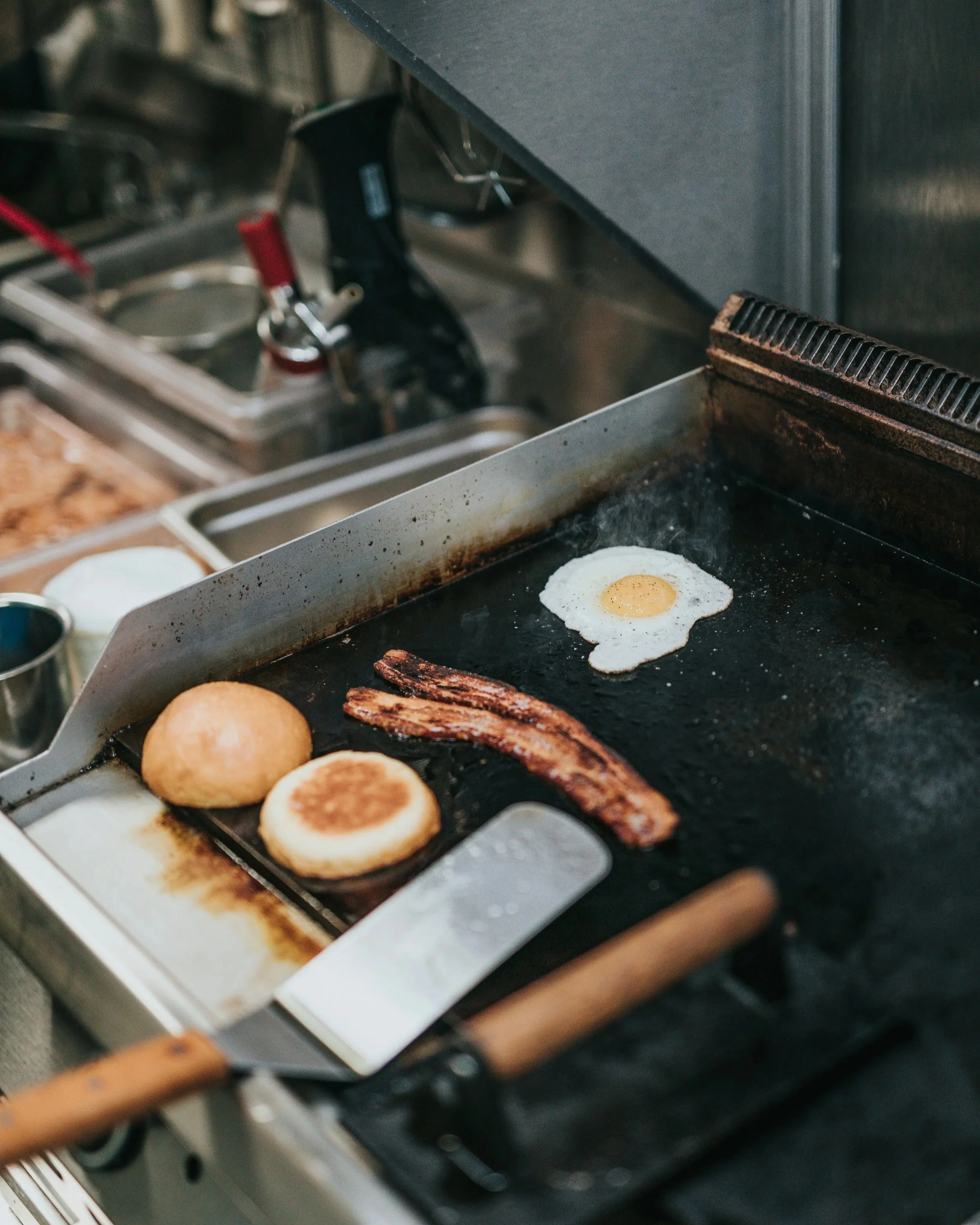 A toaster oven with three English muffins and a fried egg cooking inside, and bacon strips on a griddle.
