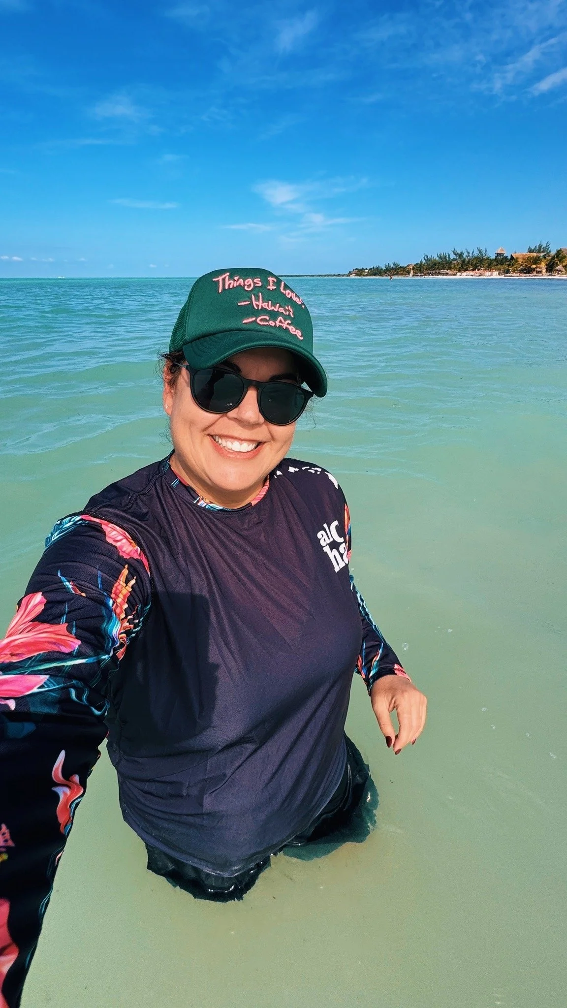 A woman smiling at the camera while standing in shallow water at the beach, wearing sunglasses, a dark floral long-sleeve shirt, a green cap, and black shorts, with a clear blue sky and distant shoreline in the background.