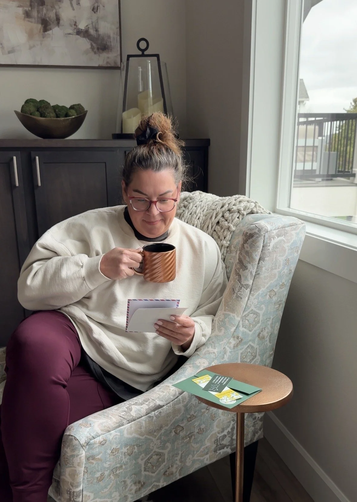 A woman with glasses and a messy bun hairstyle sitting in a light-colored armchair near a window, holding a mug and reading a card or letter. There are green promotional cards on a small round table beside her, and a dark sideboard with decorative items and a large bowl of artichokes in the background.