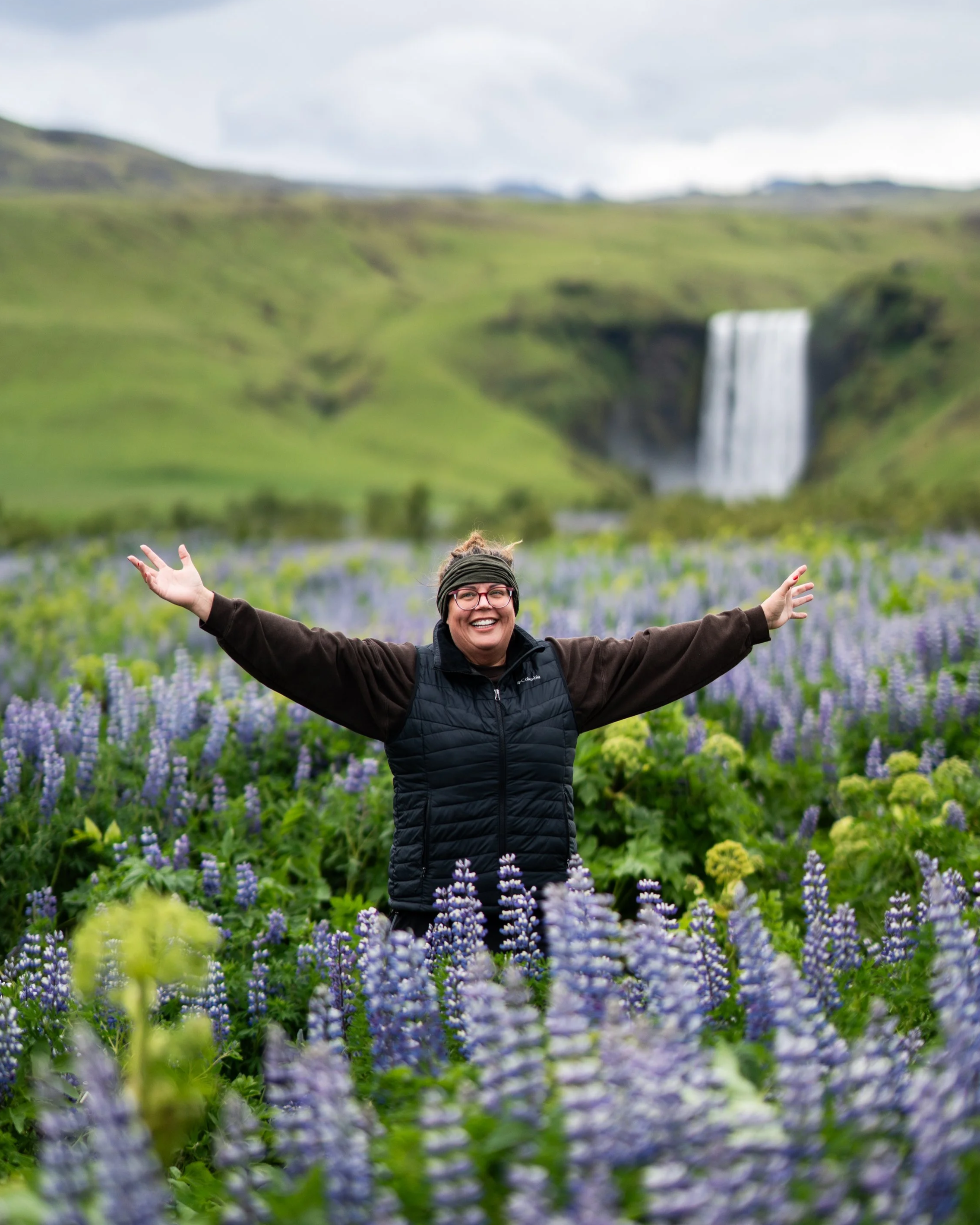 A woman smiling with her arms outstretched standing amidst lush purple and green plants, with a scenic waterfall cascading down a green hillside in the background.