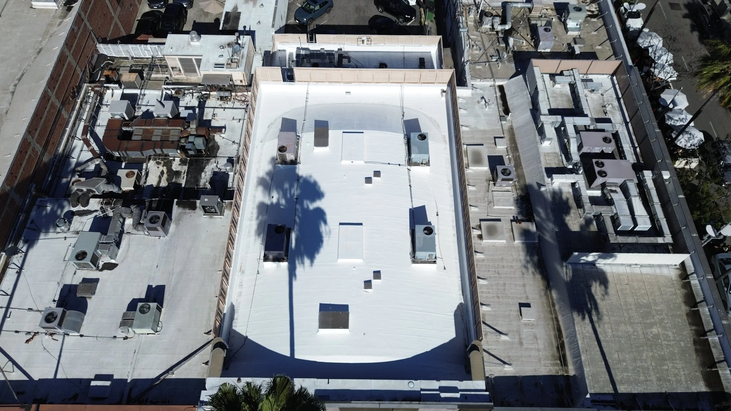 Aerial view of a building rooftop with various HVAC units, vents, and ductwork, alongside a parking lot with several cars.