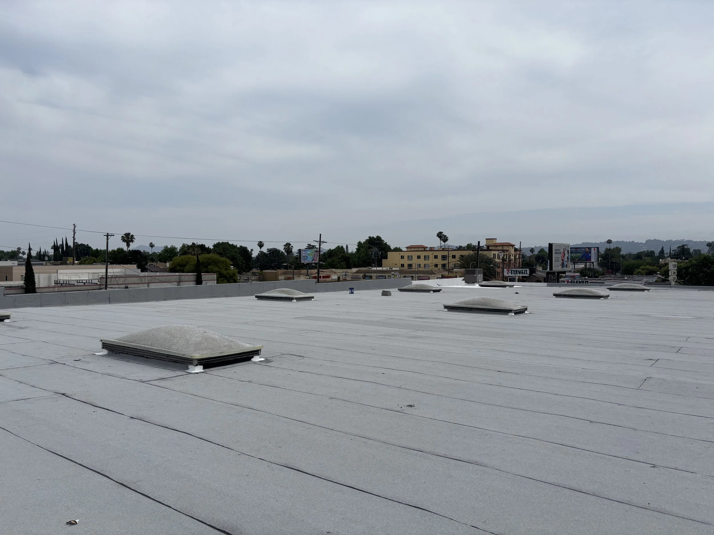 Flat commercial building roof with multiple roof vents and skylights, cityscape with trees and buildings in the background under cloudy sky.