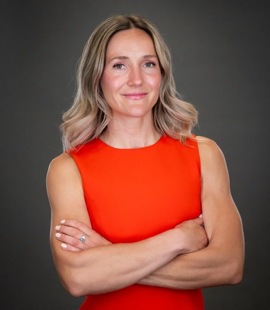 A woman with blonde wavy hair wearing a sleeveless orange dress, standing with arms crossed against a dark gray background.