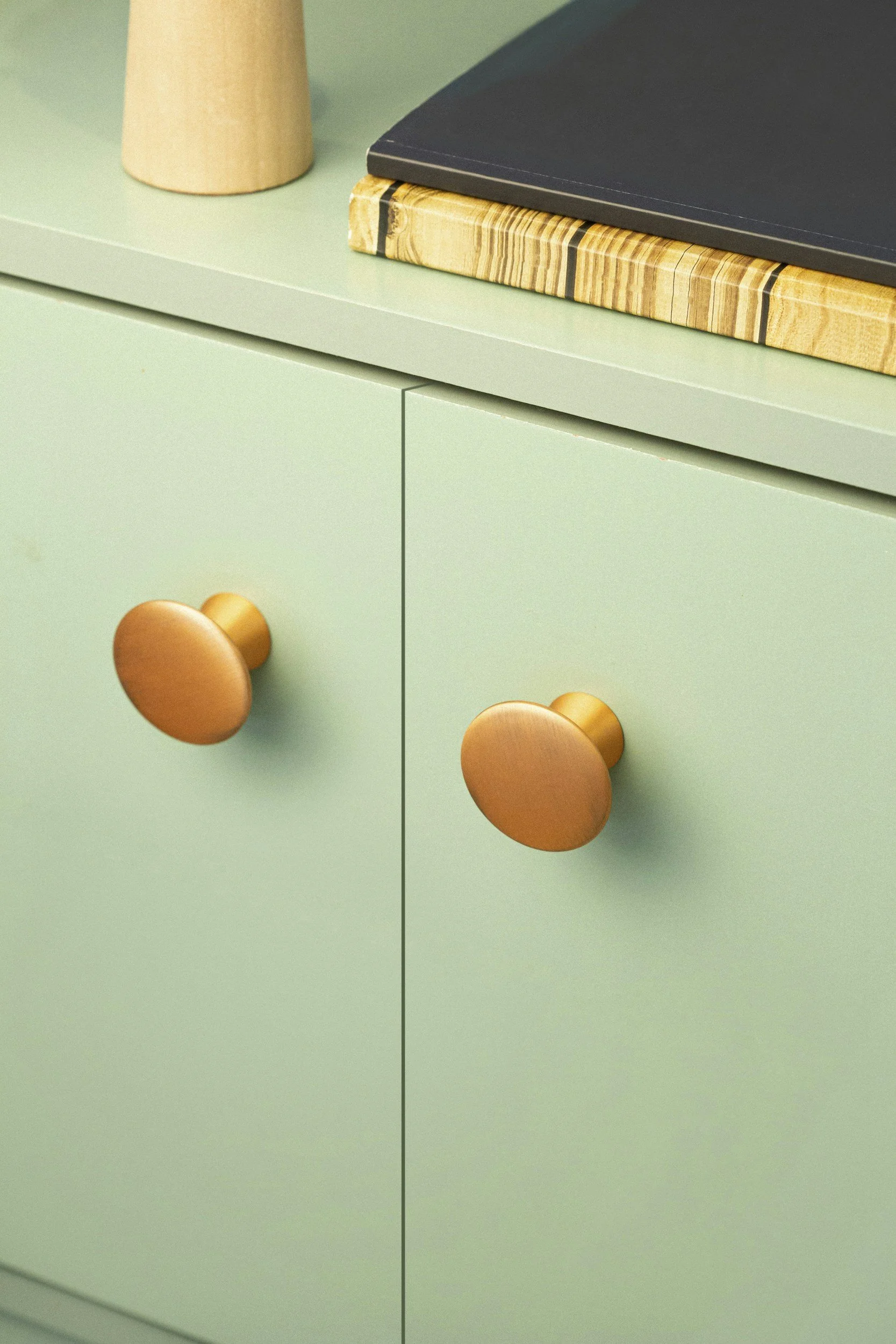 A mint green cabinet with two drawers, each with round copper-colored knobs. The top of the cabinet has a light-colored wood surface, with a black item and a stack of patterned paper or notebook placed on it. Part of a beige object is also visible on the top left corner.