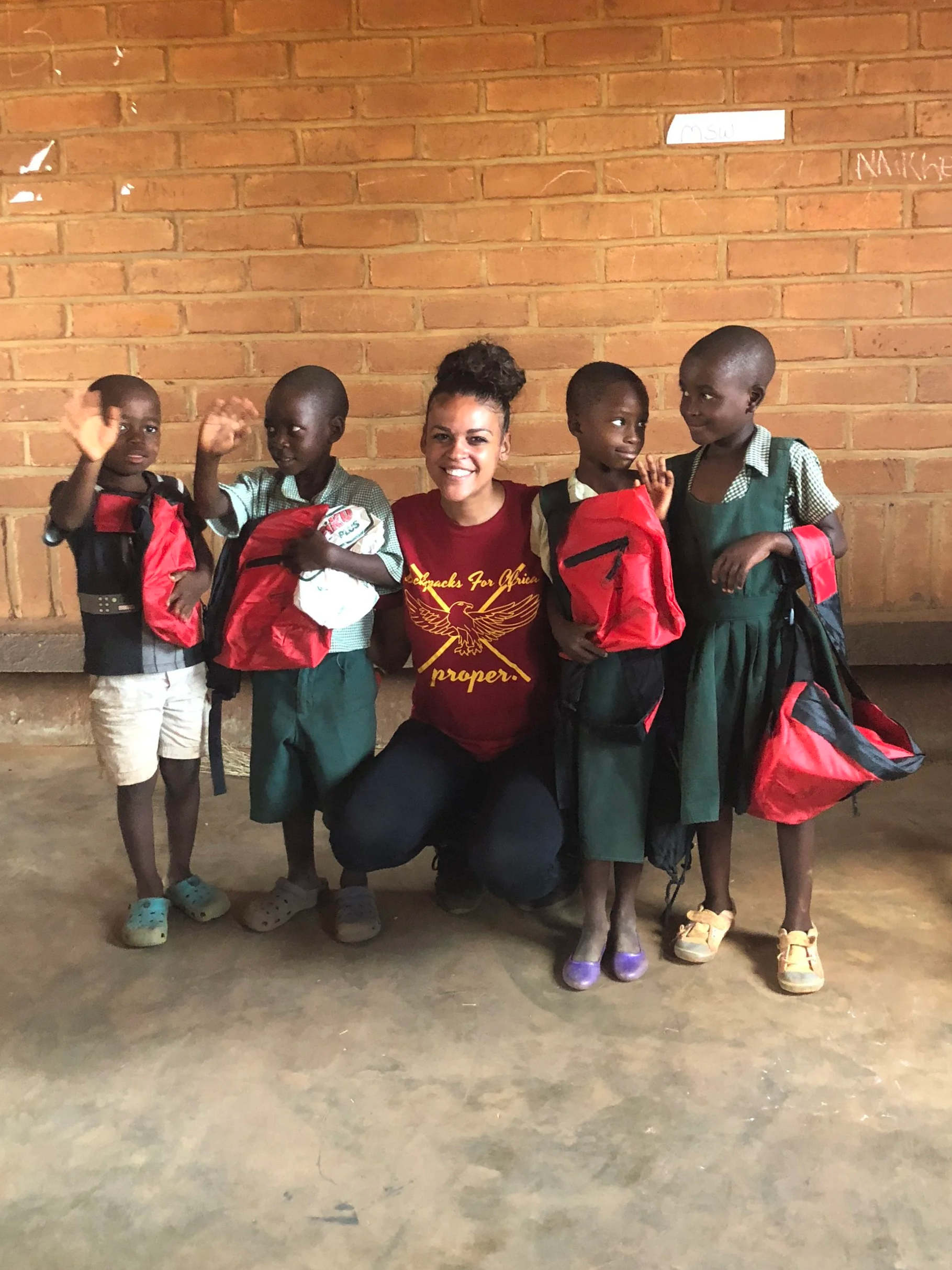 A smiling woman kneels with four young African children in school uniforms, holding backpacks, in front of a brick wall.
