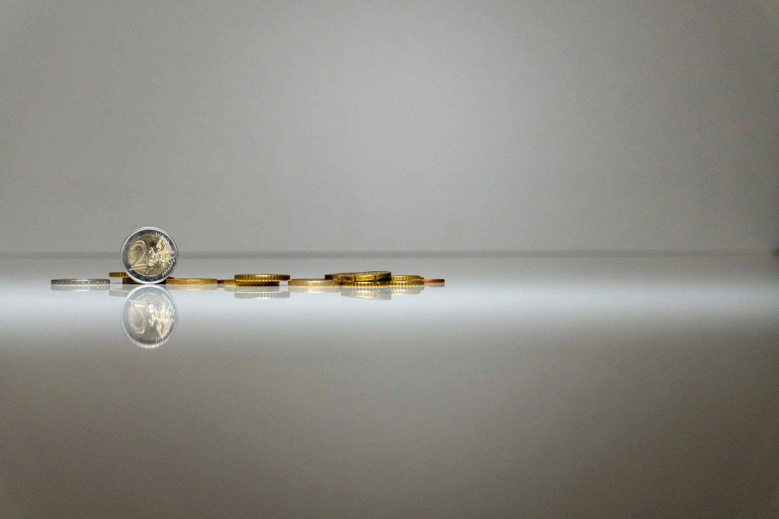 Several coins, including a two-euro coin, are lined up on a reflective surface with a plain, light-colored background.
