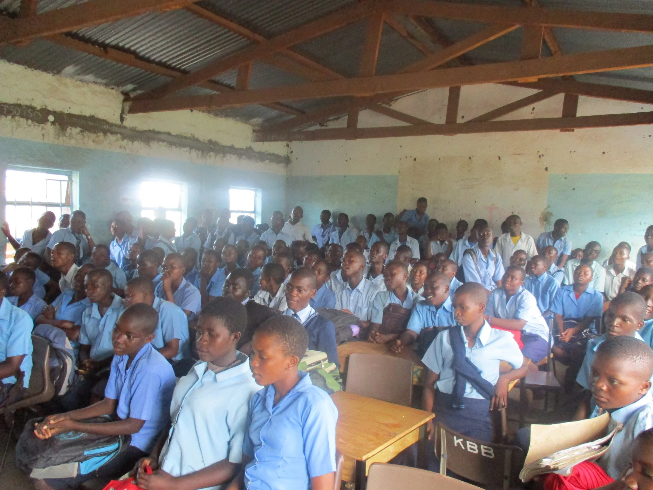 A classroom filled with students in light blue uniforms, seated and attentively listening, with sunlight streaming through windows in a modest, unfinished classroom.