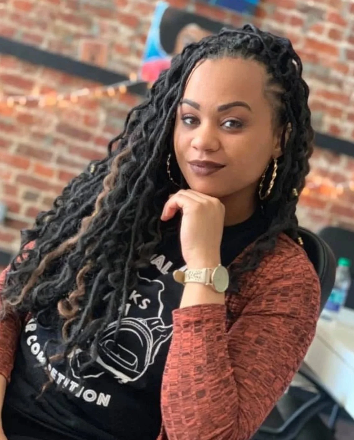 A woman with long, styled black dreadlocks, wearing a patterned orange and black top, gold hoop earrings, a beige watch, and sitting with her chin resting on her hand in an indoor setting with a brick wall background.