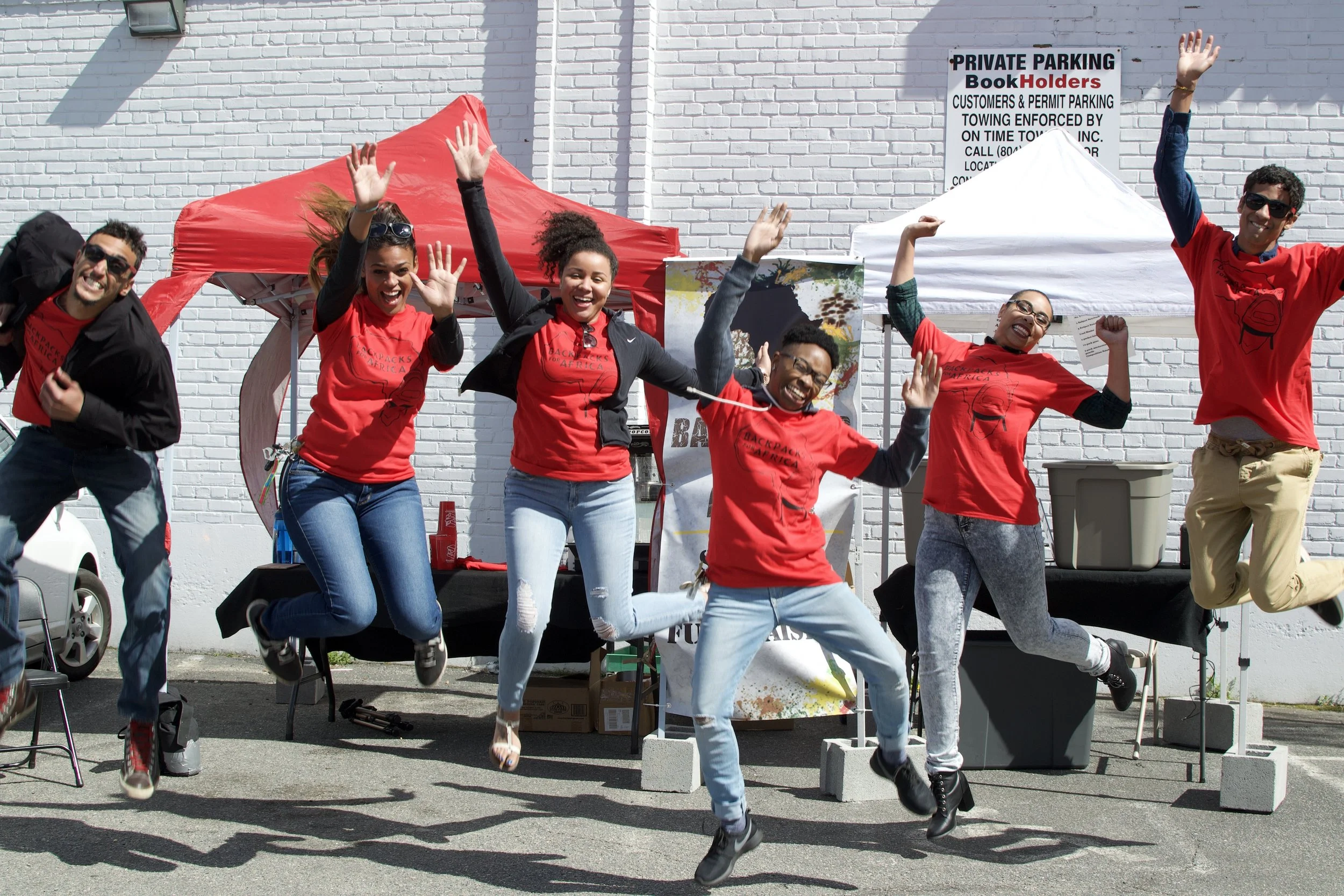 A group of six people jumping in the air and smiling at an outdoor event. They are wearing red shirts, and there are tents and a sign in the background.