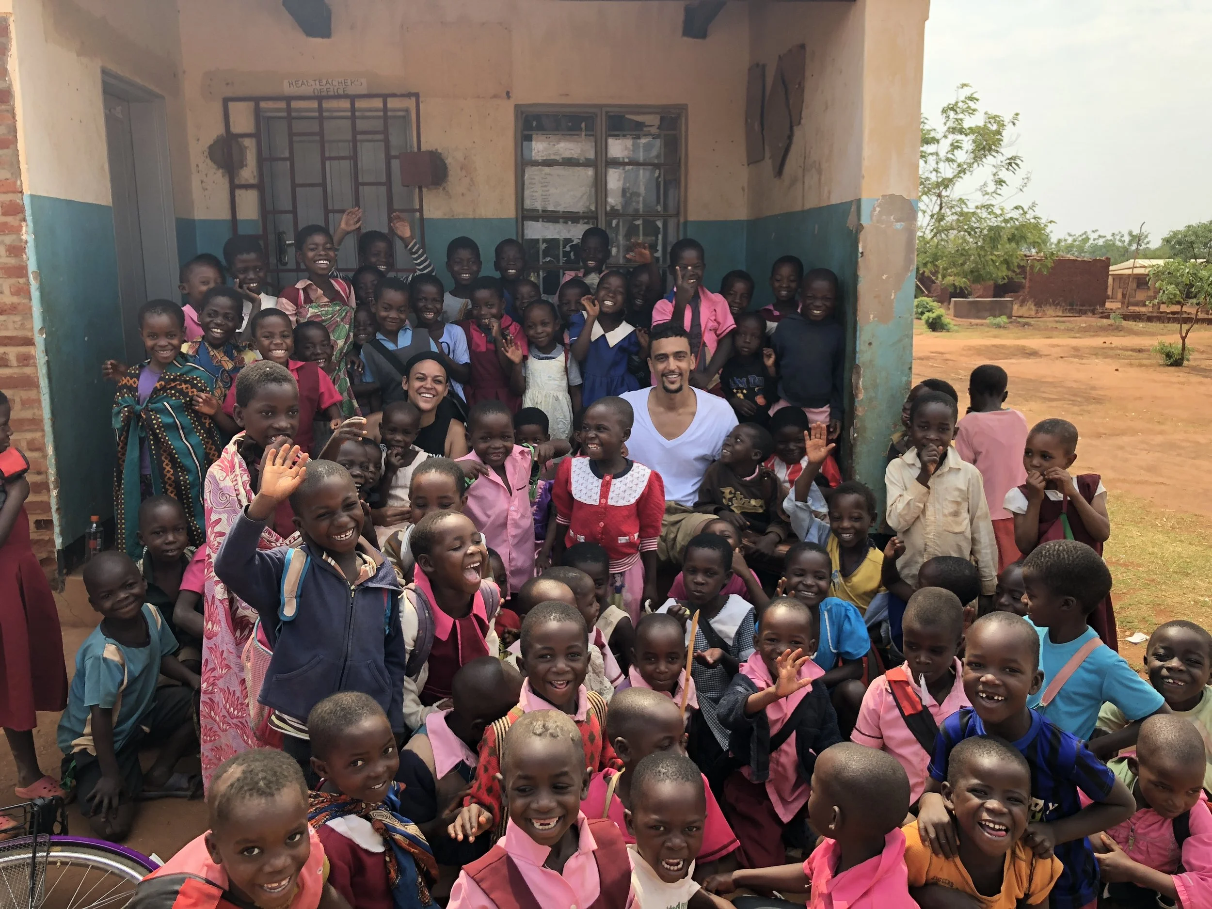 Group of smiling children and two adults posing outside a building with weathered walls and a blue-green lower wall, in a rural setting with dirt ground and sparse trees.