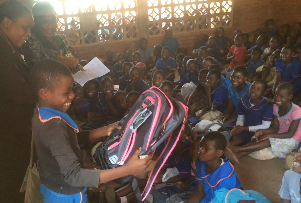 A young boy in school uniform is smiling as he receives a backpack from an adult woman during a school event. In the background, many children are seated on the floor, smiling and watching the exchange, inside a large room with wooden lattice windows.