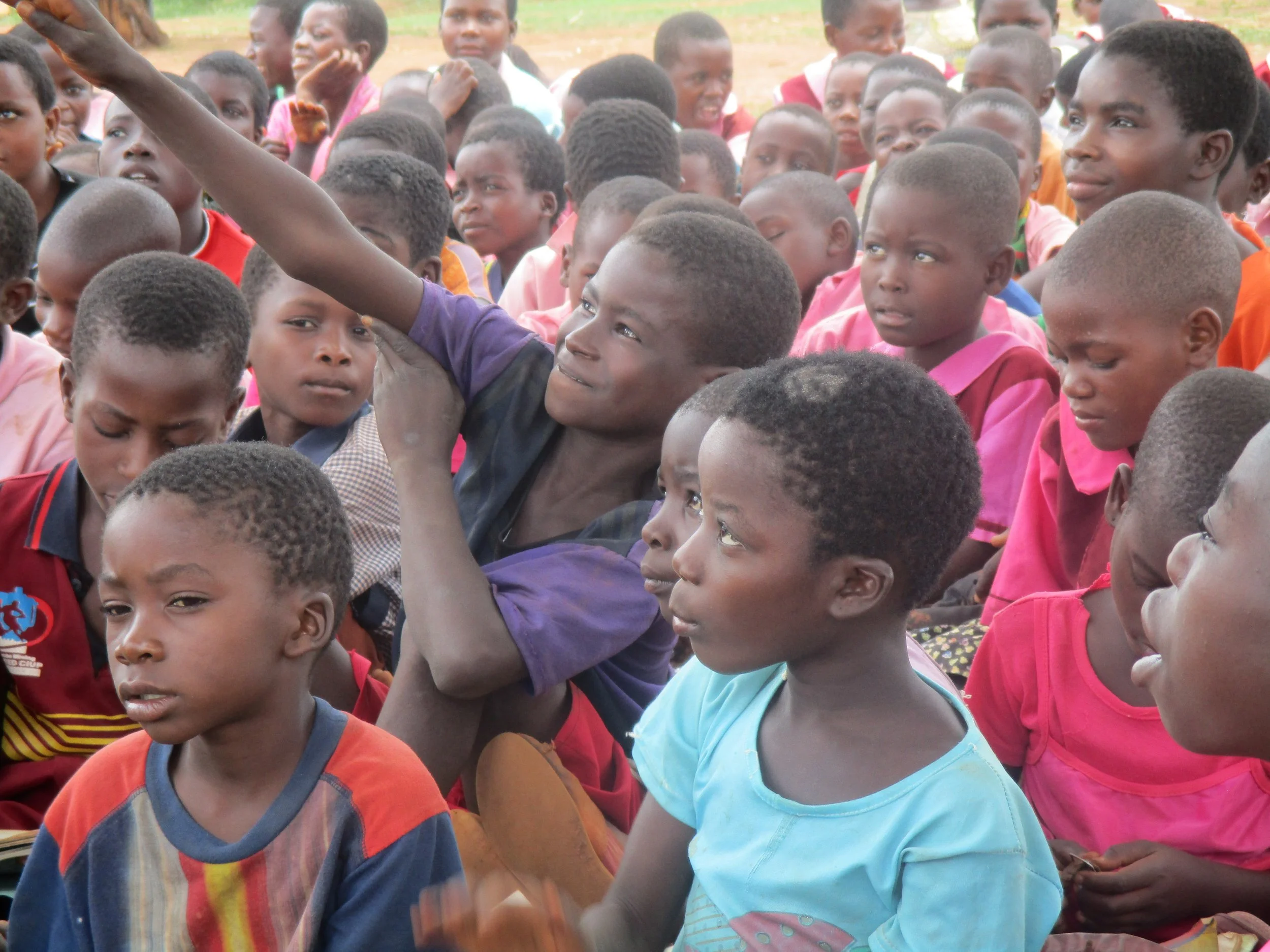 A group of children sitting outdoors, attentively listening or watching something, with one boy raising his hand.