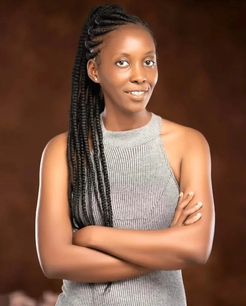 Portrait of a woman with braided hair, arms crossed, wearing a sleeveless striped top, smiling slightly, against a brown background.