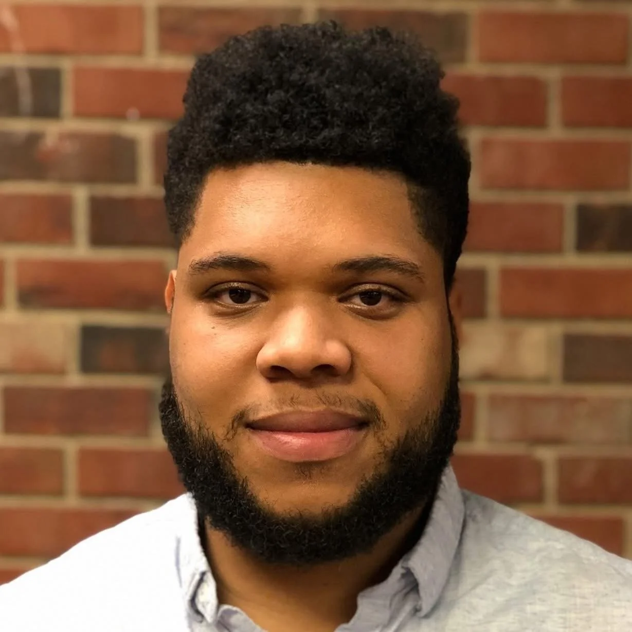 A young man with a short curly hairstyle and a full beard, wearing a light gray collared shirt, standing in front of a red brick wall.