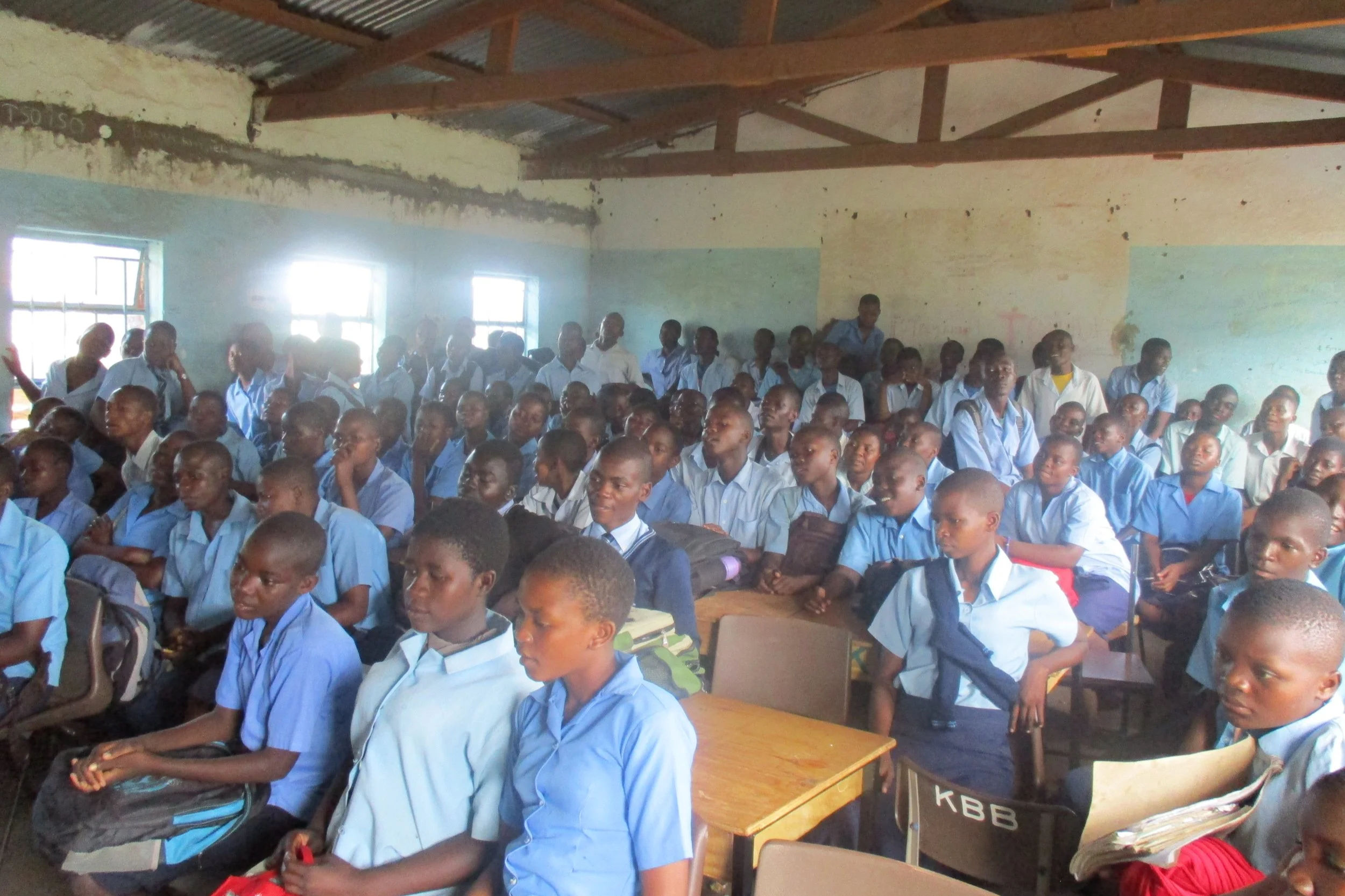 A classroom filled with numerous students wearing light blue school uniforms. The students are seated and some are standing, facing forward. The classroom has light coming in through the windows, and the walls are painted with blue and beige sections. The room has a high, corrugated metal roof with wooden beams.