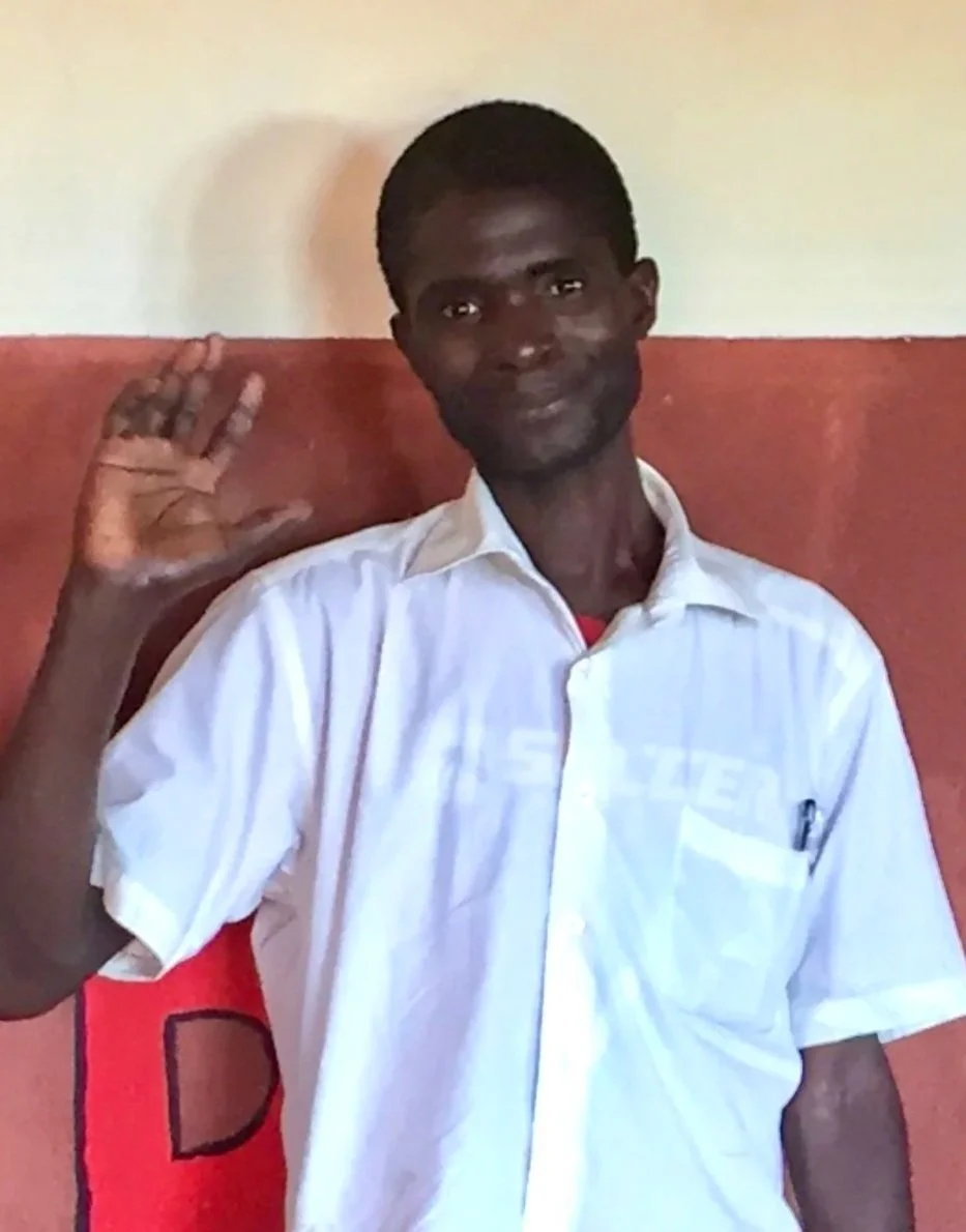 A young man in a white shirt waving, standing in front of a red and cream wall.