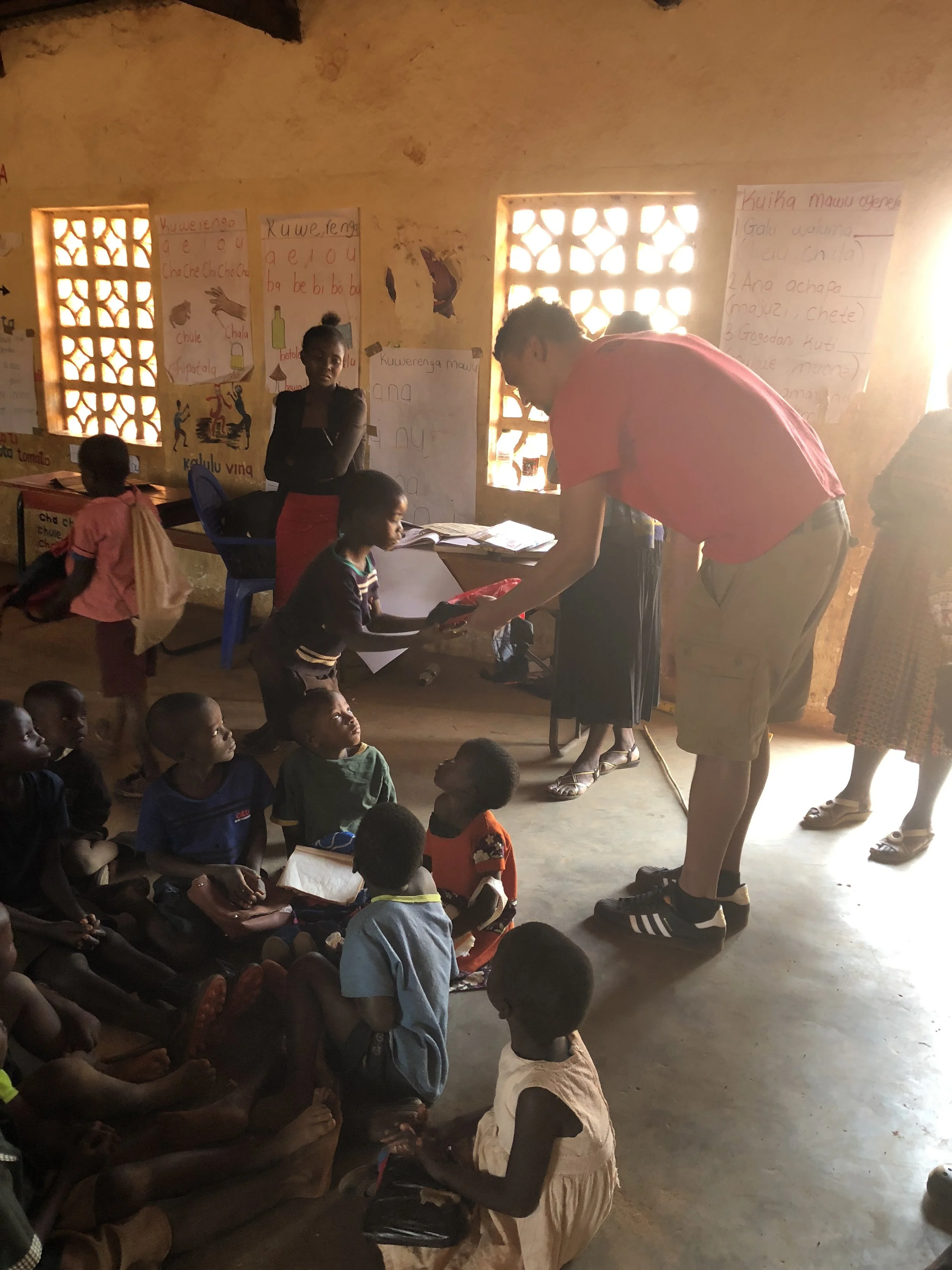 A teacher hands a book to a young girl in a classroom with children seated on the floor. The classroom walls have educational posters, and several children watch attentively.