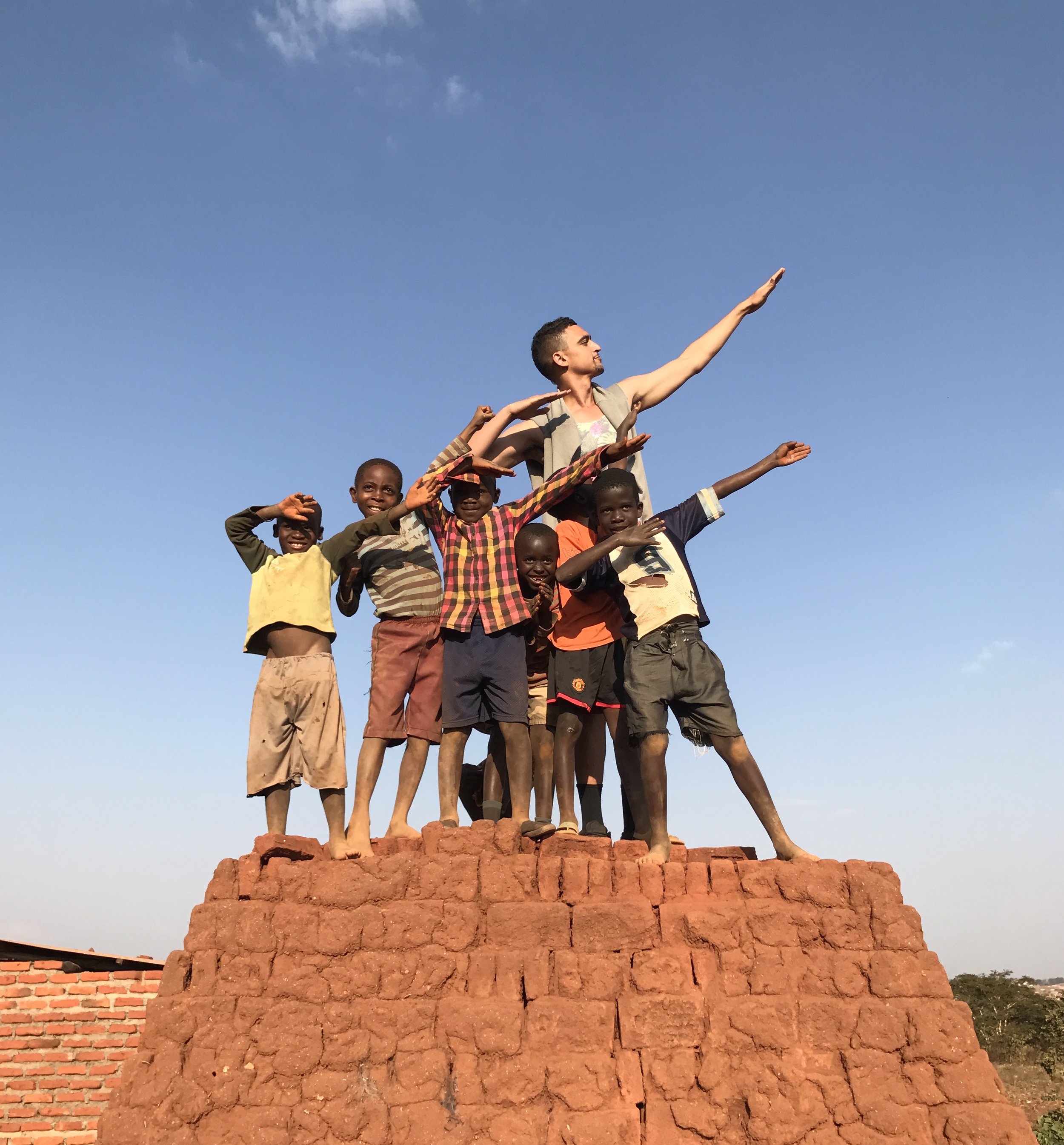A group of children and an adult standing on a brick platform outdoors, striking a pose with arms extended in a victory salute against a clear blue sky.