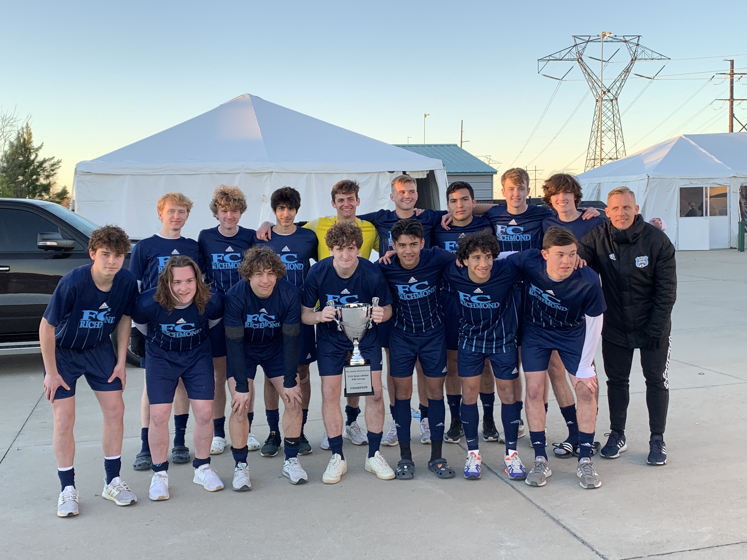 A youth soccer team in navy blue jerseys celebrating a victory, posing with a trophy in an outdoor area during sunset, with tents and power lines in the background.