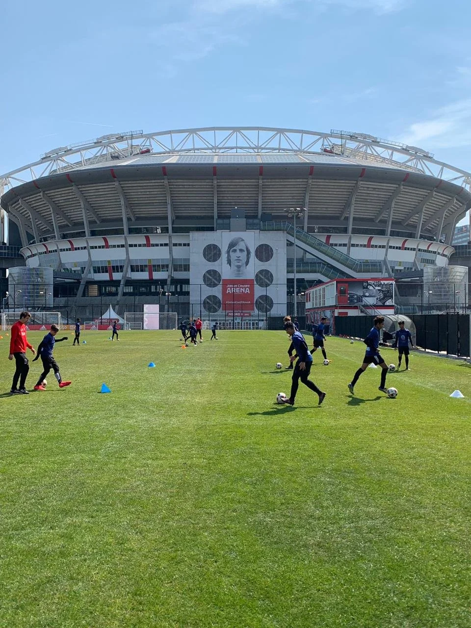 Young soccer players training on a green field outside the John Crusoe Arena stadium with a large image of a person and a modern, circular stadium in the background.