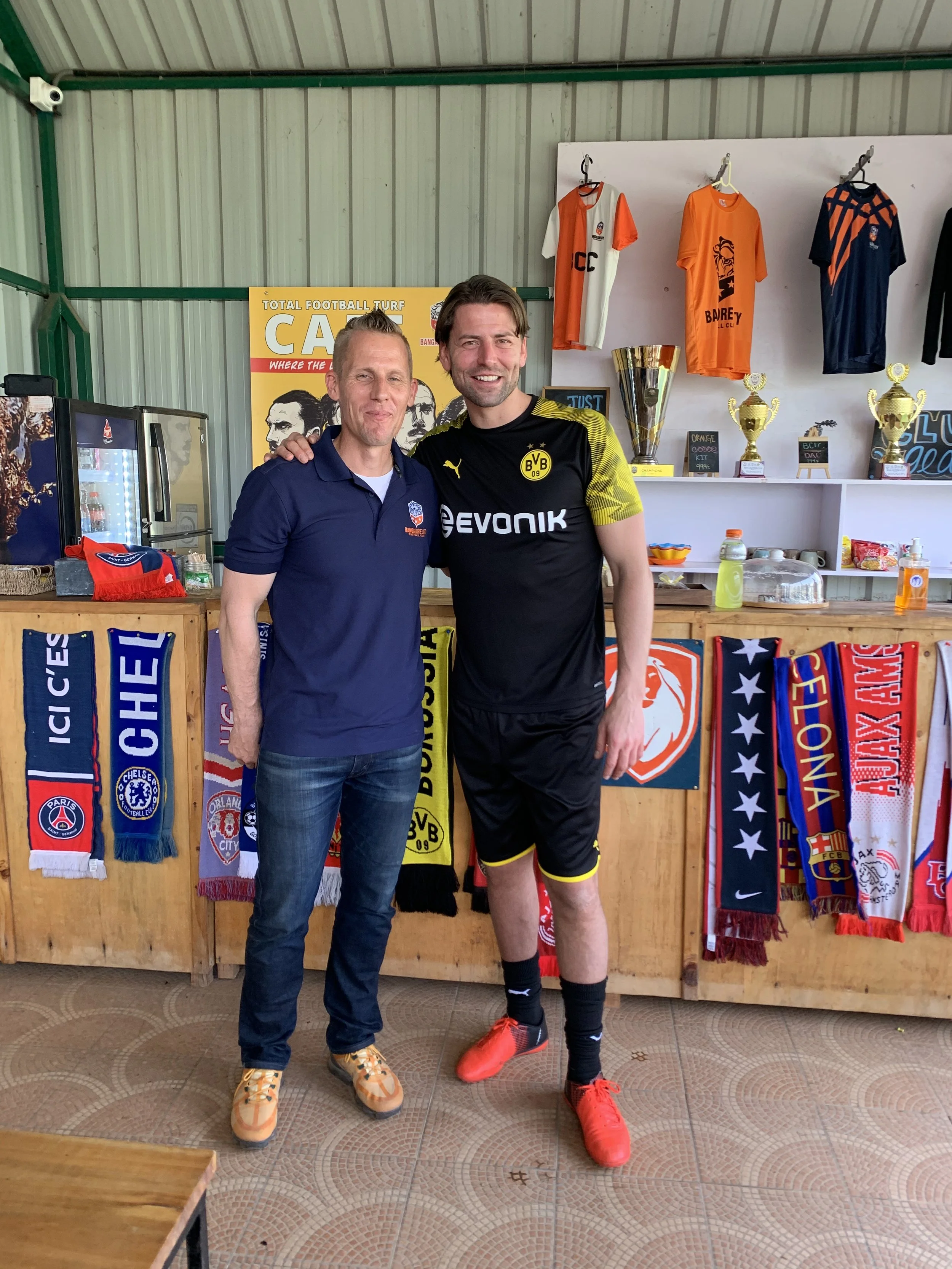 Two men standing together in a sports-themed room, smiling at the camera. One is wearing casual clothes and the other is in a football uniform. Behind them are sports jerseys, trophies, and scarves from various football teams.