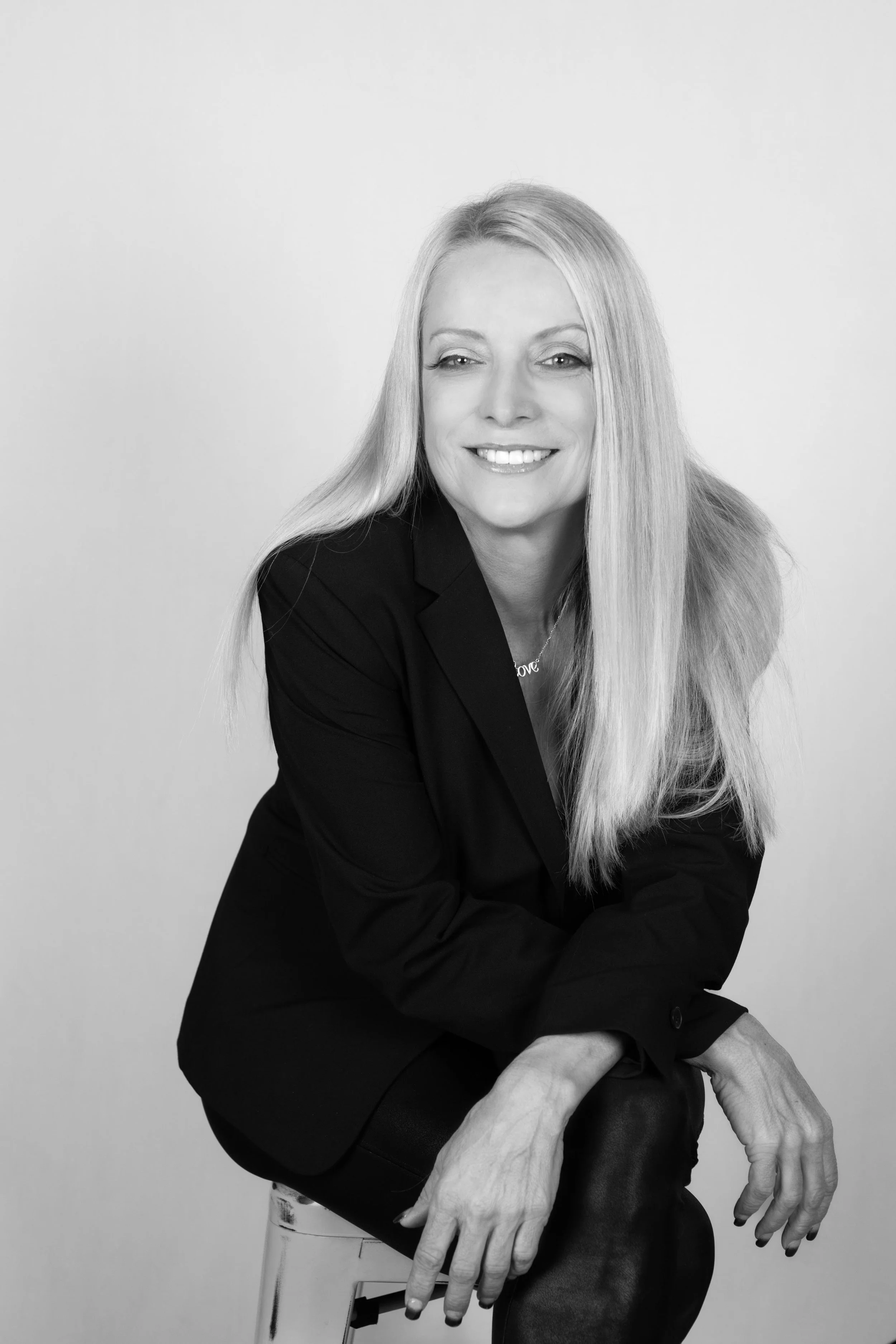 Black and white photo of a woman with long, straight blonde hair wearing a light-colored top, standing in front of a wall with a tree branch painted on it.