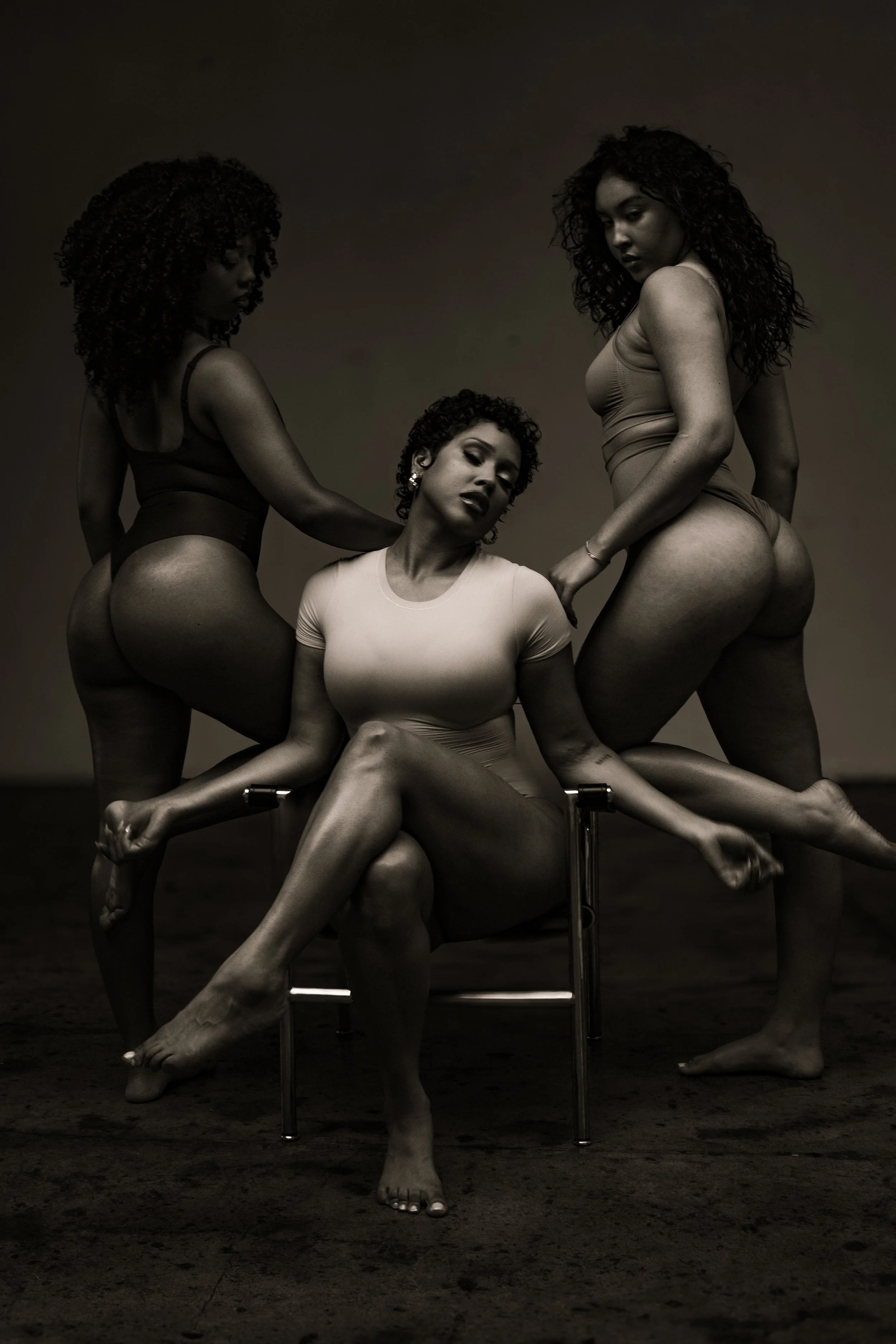 Black and white photo of three women with curly hair posing together, one sitting, two standing, in a studio setting with dark background.