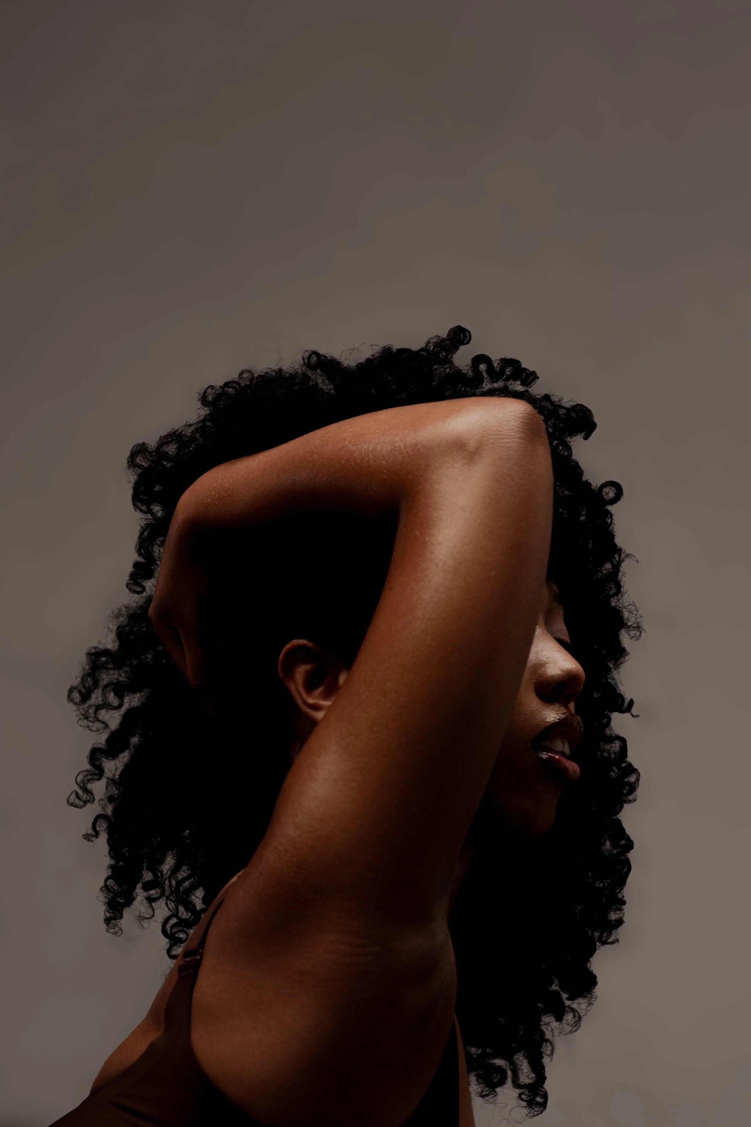Close-up of a woman with natural curly hair, with her arm raised over her head, partially covering her face, against a neutral background.