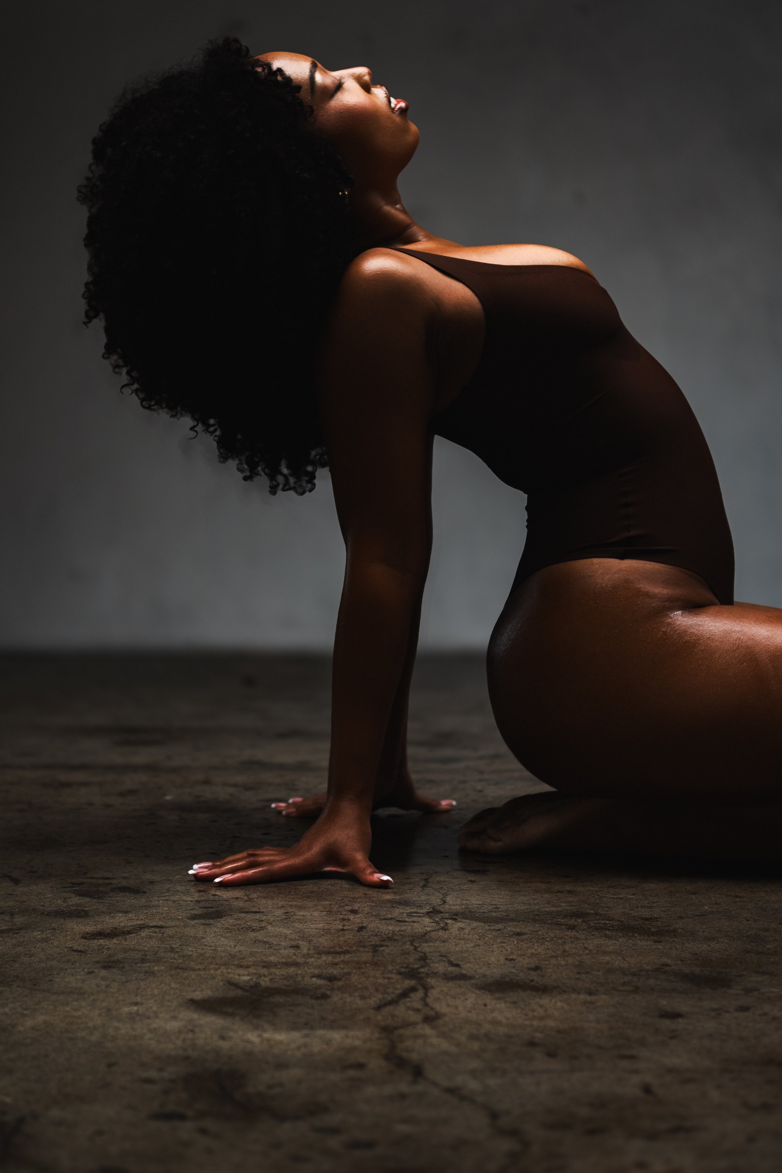 A woman with curly black hair kneels on a dark, textured floor with her head tilted back, eyes closed, and a serene expression. She is wearing a black sleeveless top, and her body is illuminated with soft lighting against a plain, dark background.