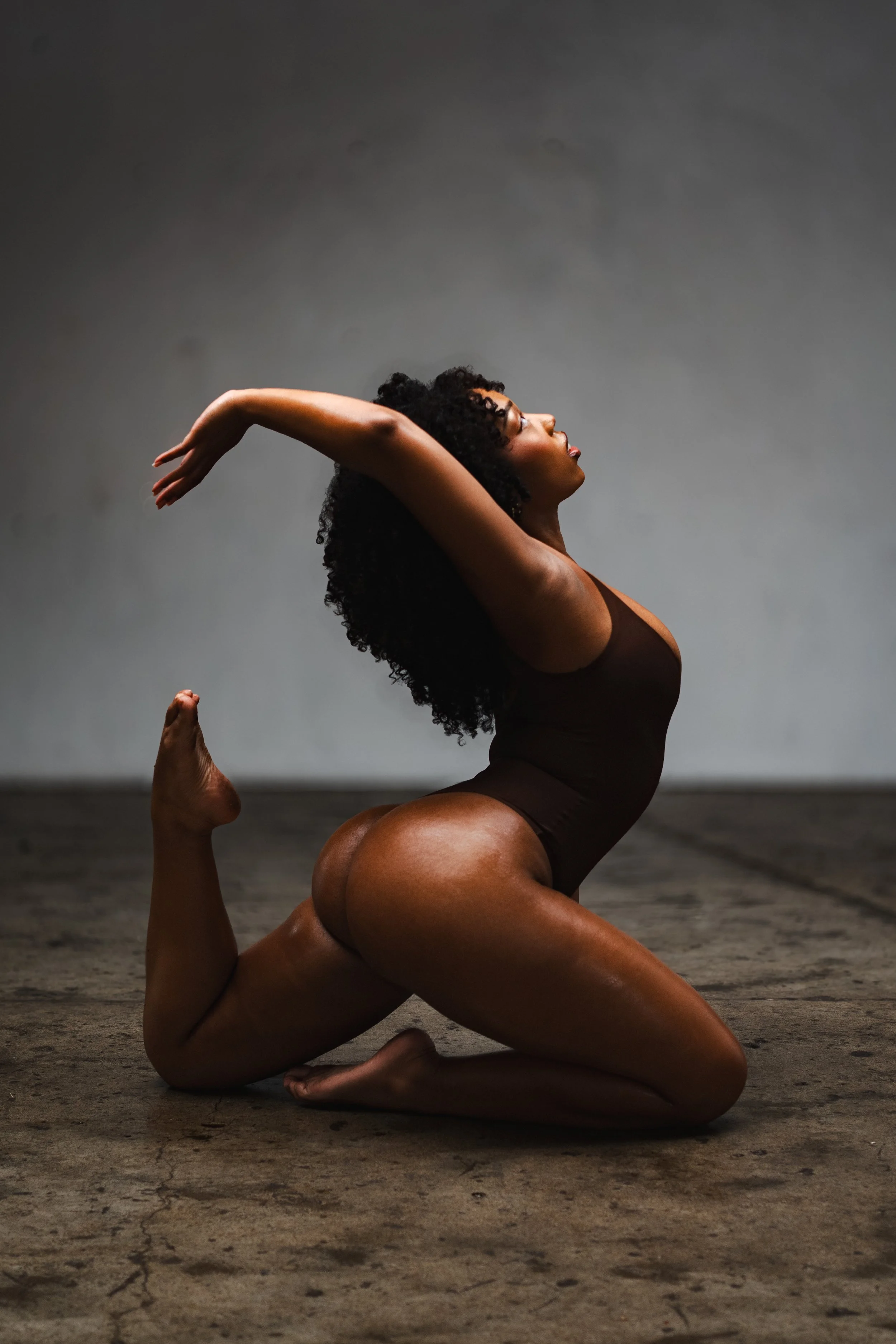 A woman with curly hair doing a yoga pose on a dark, textured floor against a plain gray background. She is on her knees with her arms extended back and her head tilted upward.
