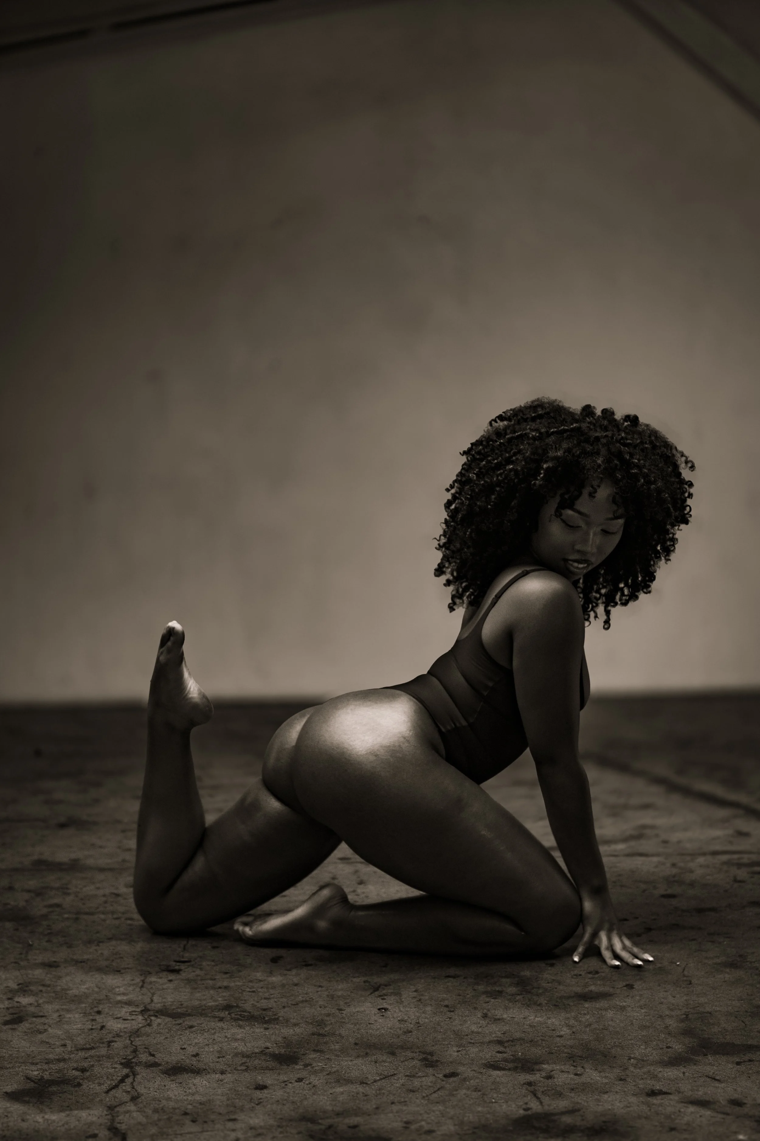 A woman with curly hair performing a dance pose on a wooden floor in a dimly lit studio.