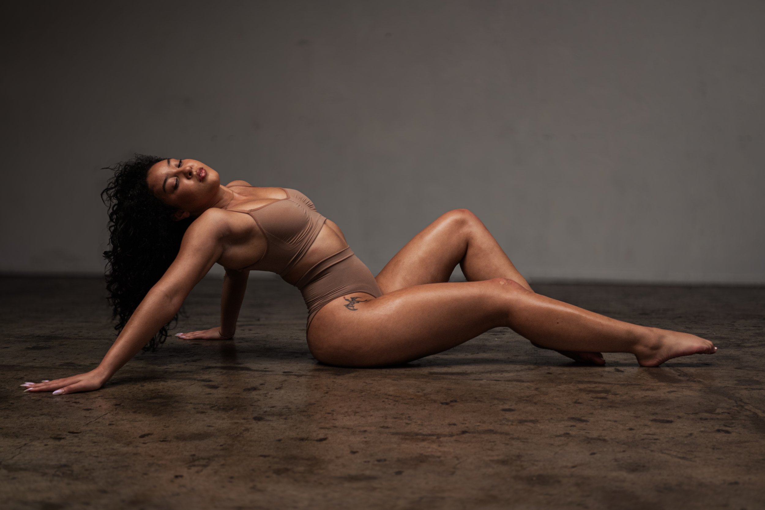 A woman with dark curly hair in a tan sports bra and shorts poses on a dark wooden floor against a gray background, sitting with her back arched and her eyes closed