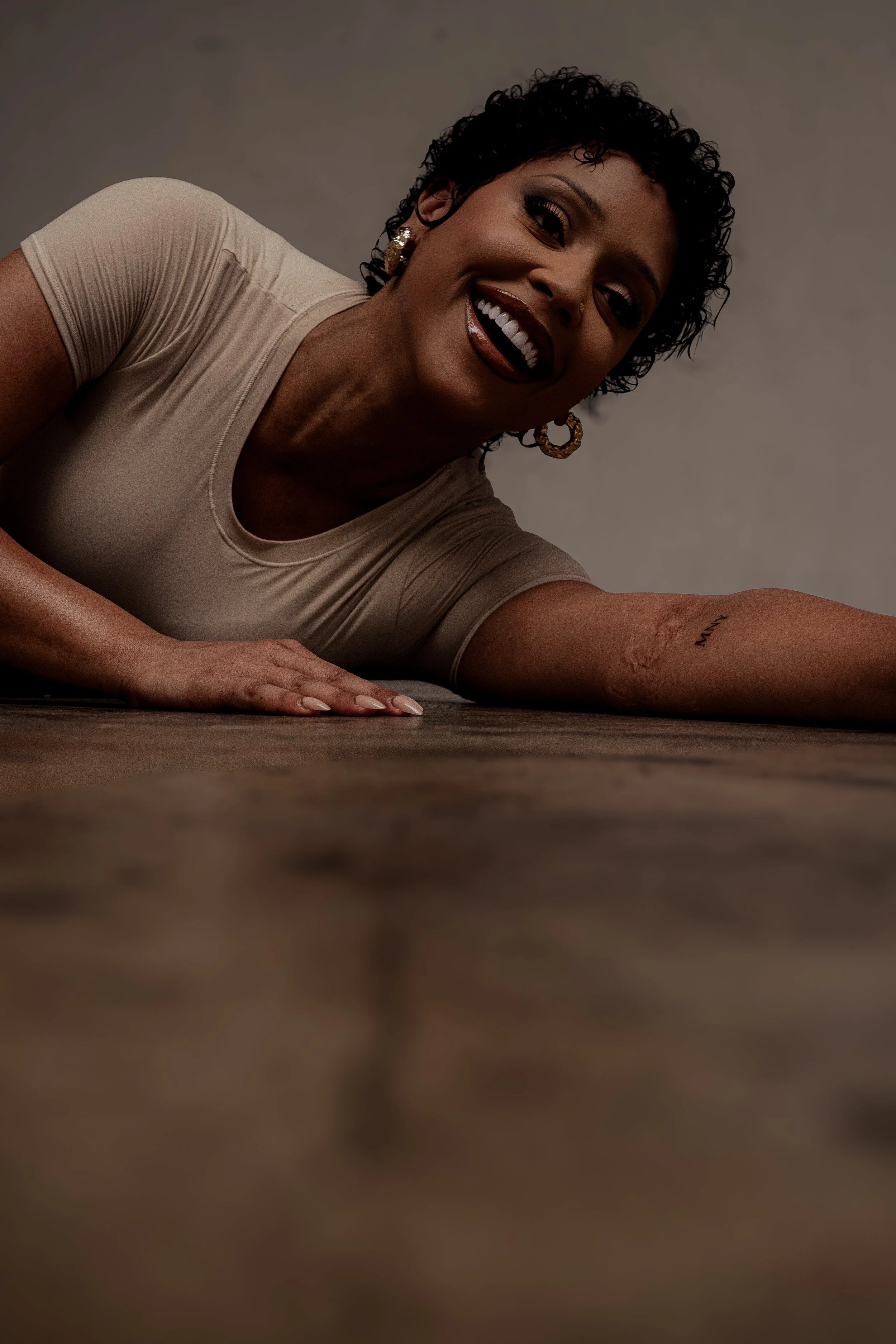 A woman with dark curly hair and earrings smiling, lying on the floor with her face close to the camera.