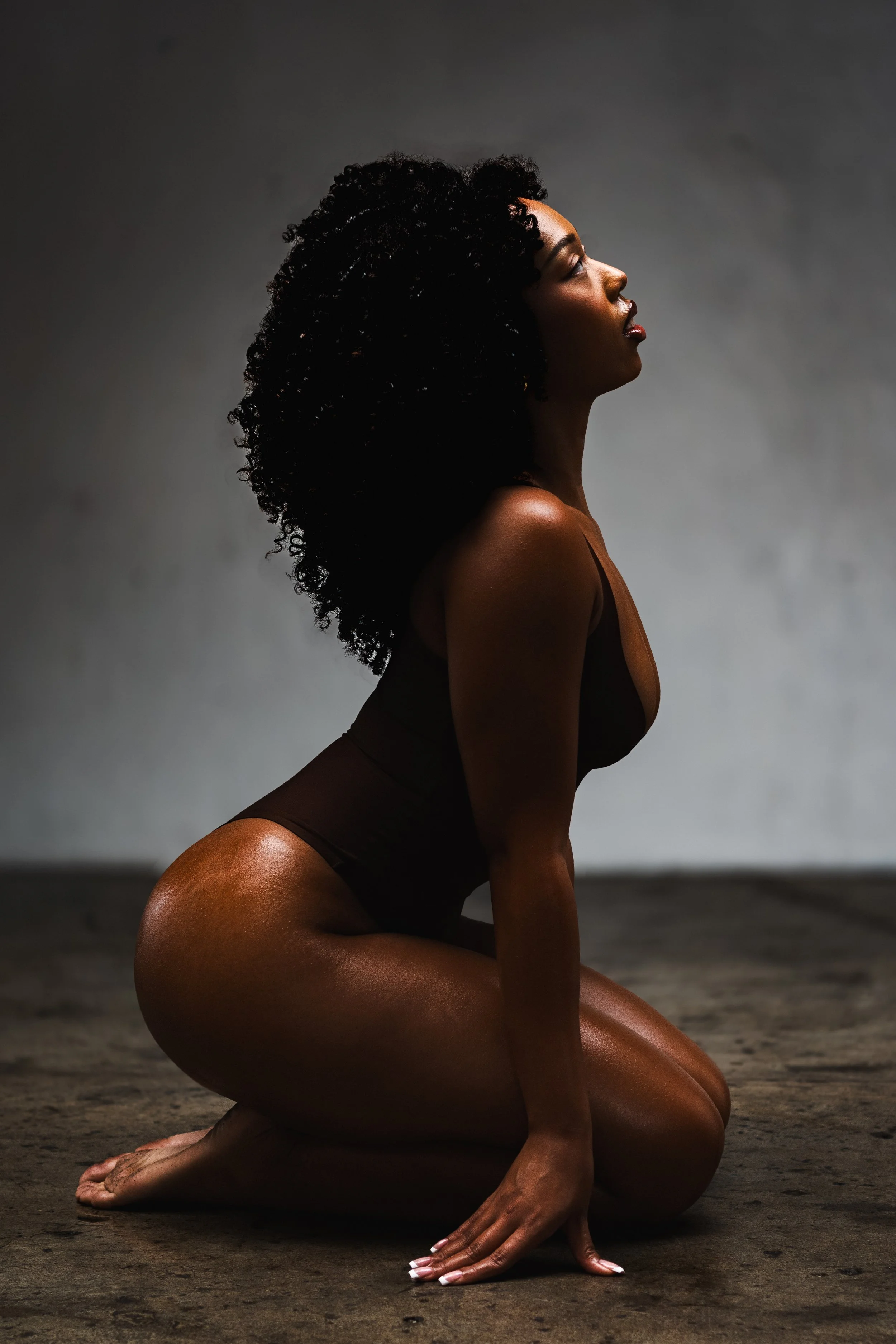 A woman with curly black hair crouches on a dark ground, profile view, with a simple gray background.