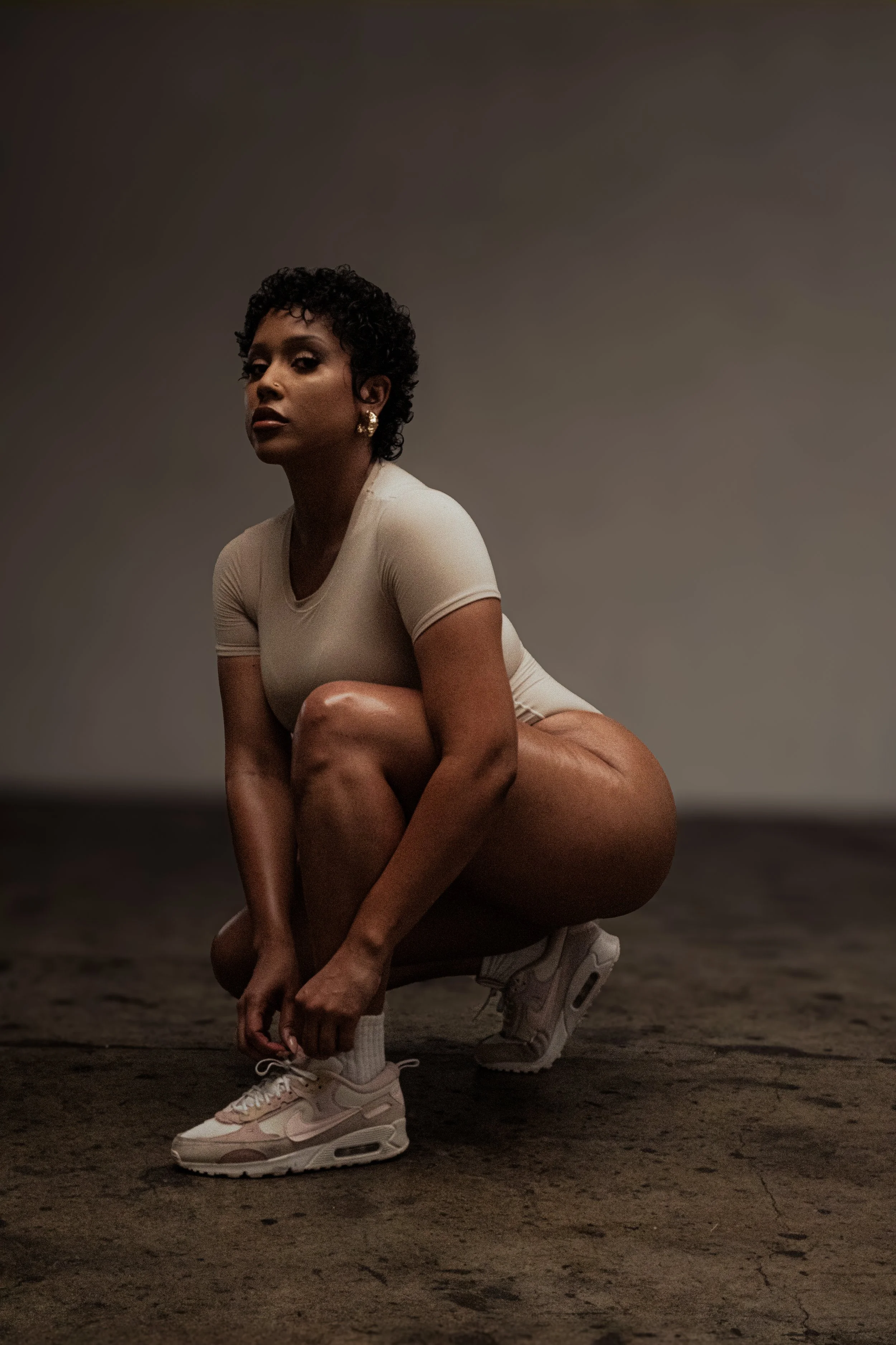A woman with short, curly hair is squatting on the ground, tying her shoelaces, wearing a beige t-shirt, white sneakers, and large earrings, against a plain gray background.