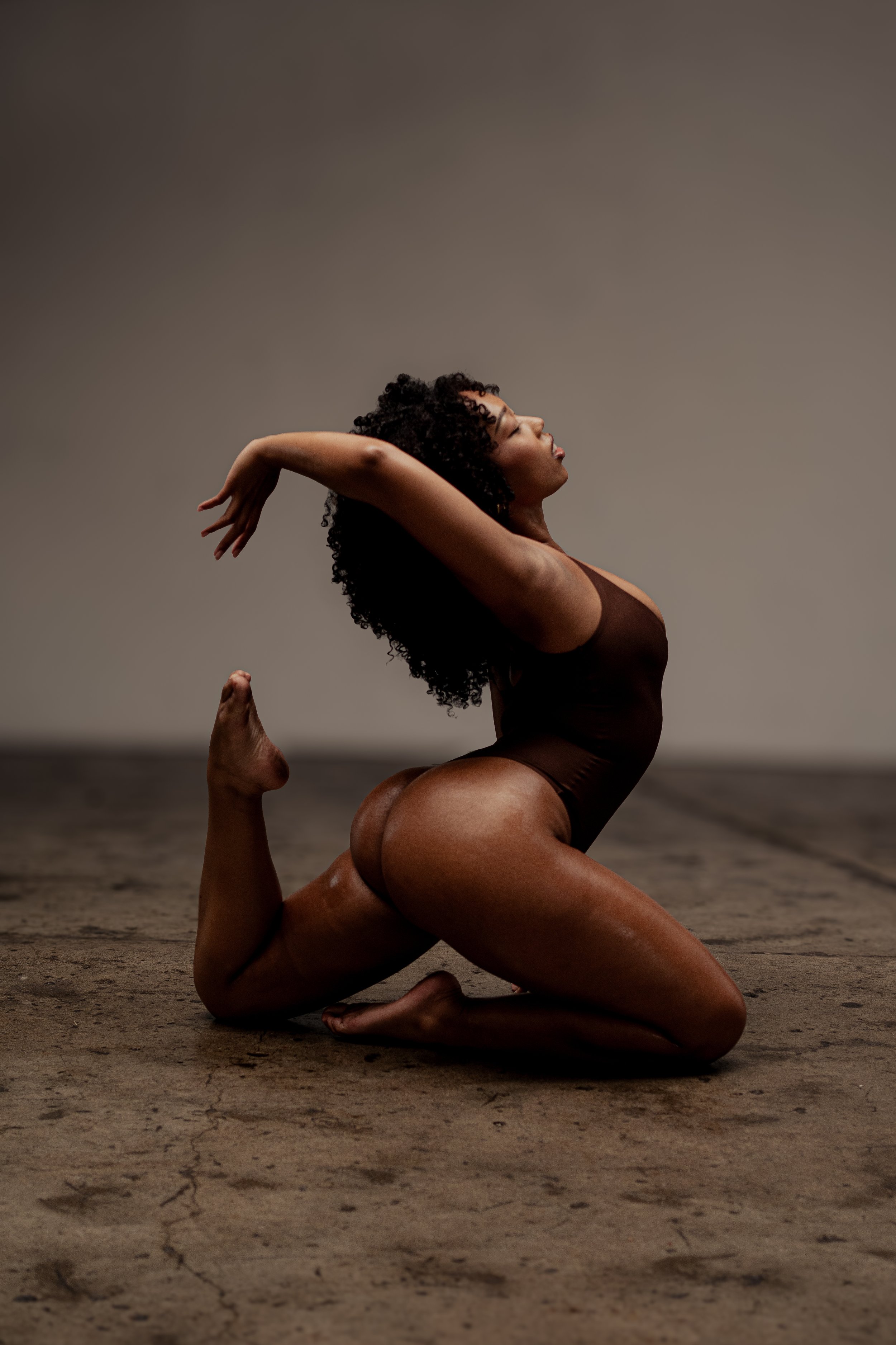 A woman with curly hair practicing yoga on a textured ground in a low, kneeling pose with her arms extended backward and her head tilted slightly upward against a plain gray background.