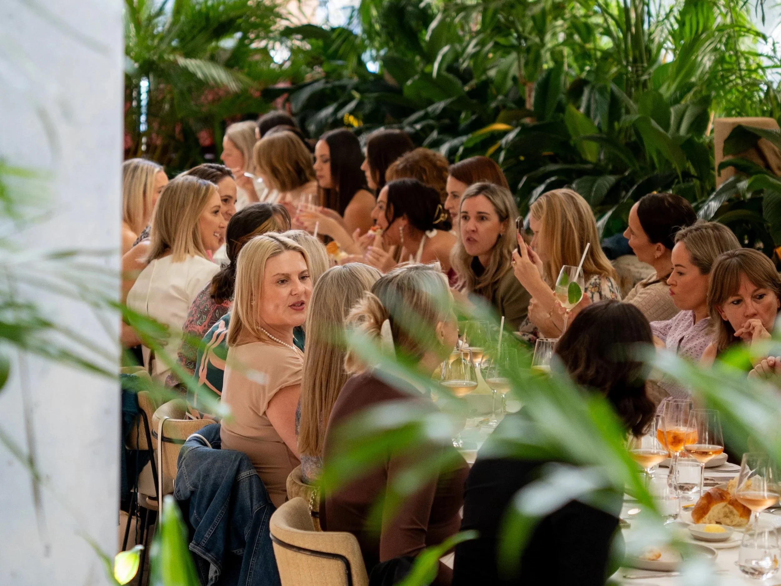 A group of women sitting at a long table during a social gathering or meal, surrounded by greenery.