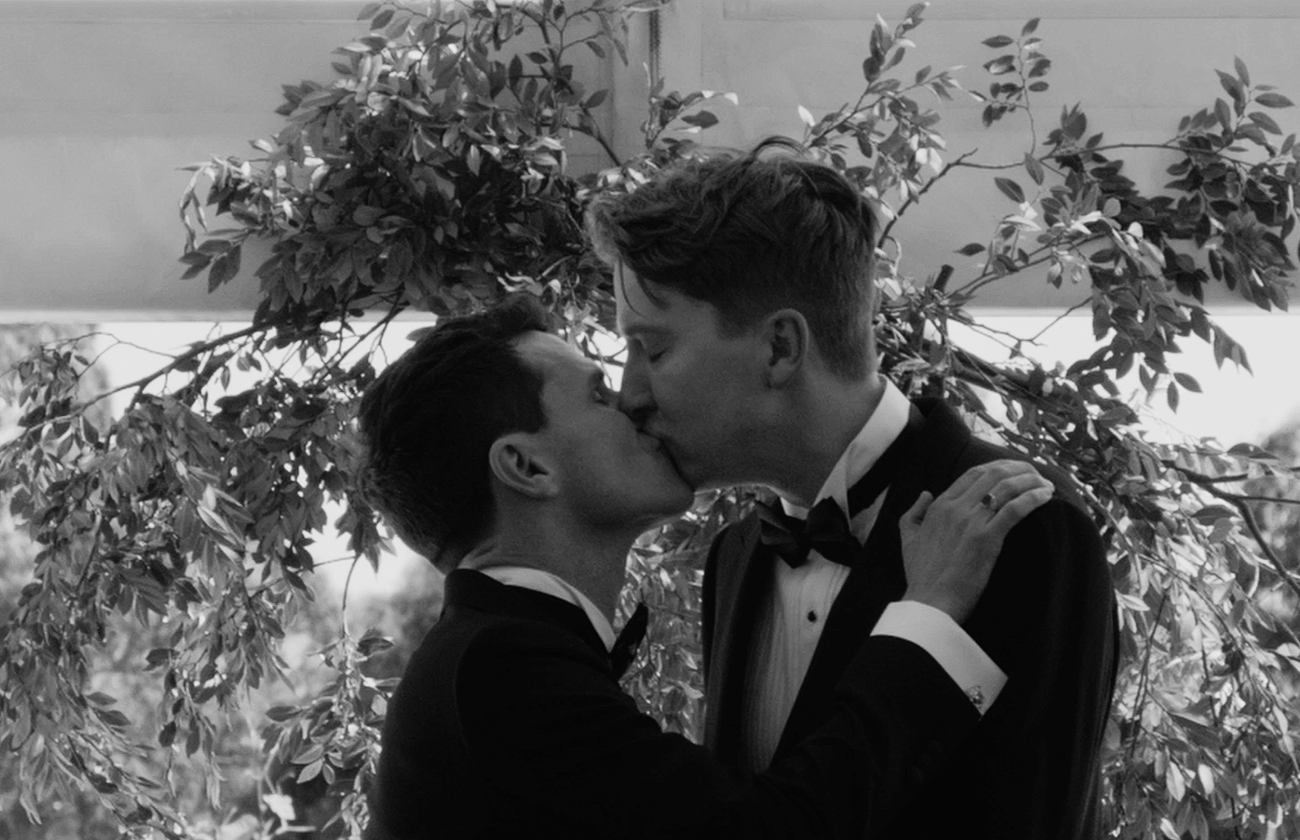 Two men in tuxedos sharing a kiss in front of a leafy background, with one man holding the other by the shoulders, during a formal event.