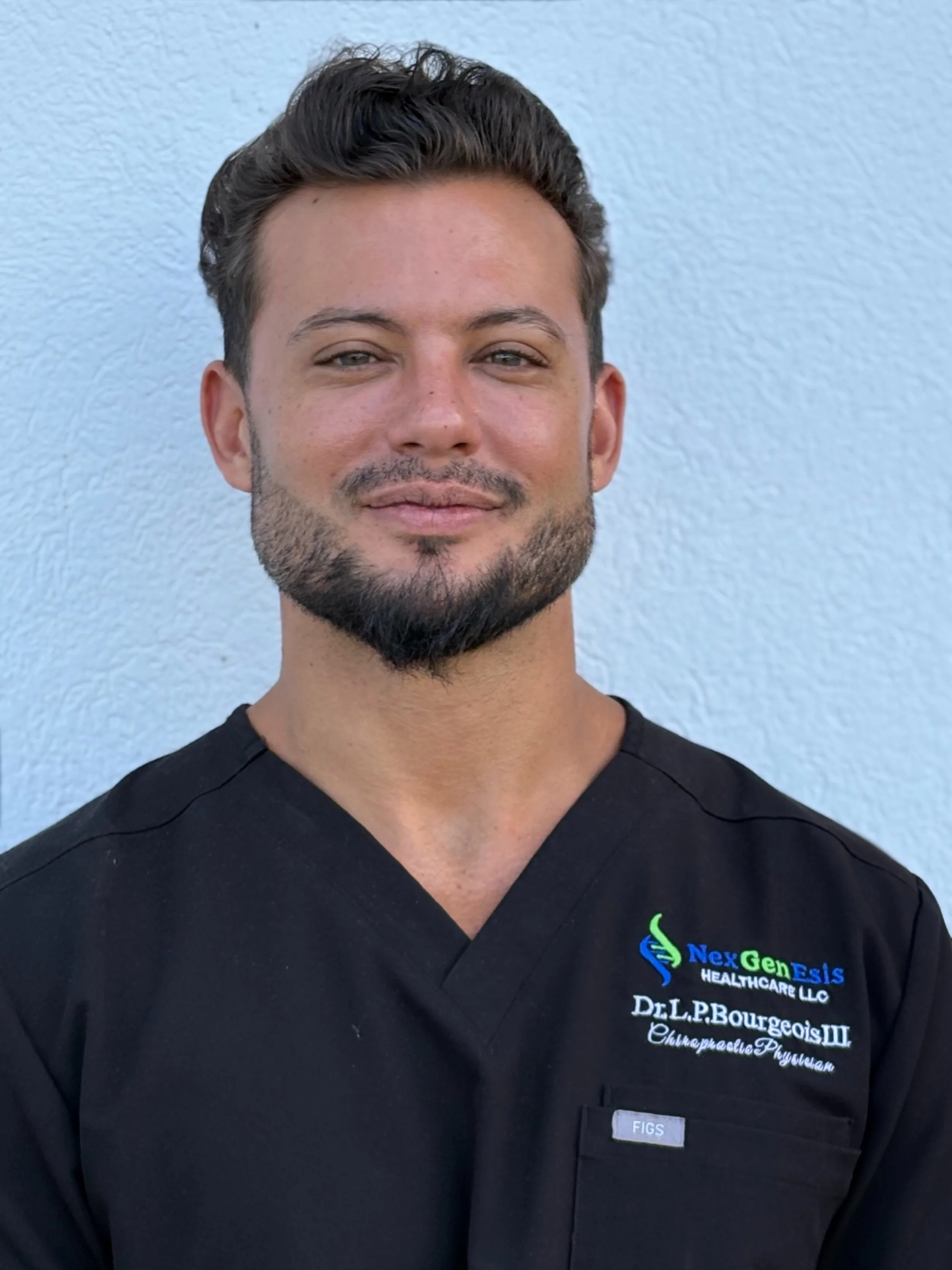 A man with dark, styled hair and a beard, wearing a black medical uniform with branding for NexGenEIs Healthcare and Dr. L.P. Bourgeois III, Chiropractor Physician, standing in front of a light-colored wall.